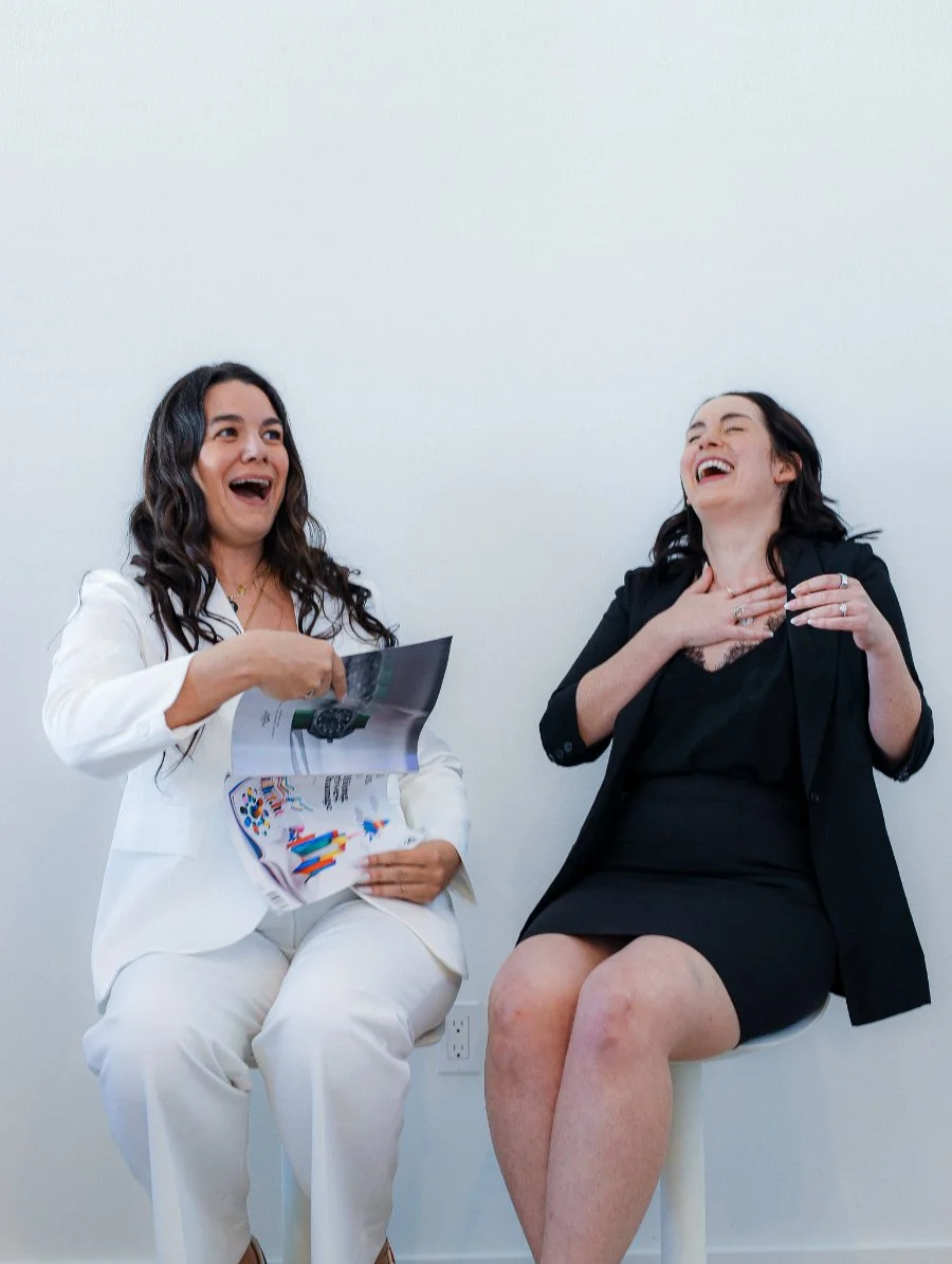 Two women sitting side by side laughing and reacting to something they’re reading together, holding printed materials. The candid energy captures collaboration, connection, and the kind of real-life moments behind a thoughtful summer sales strategy.