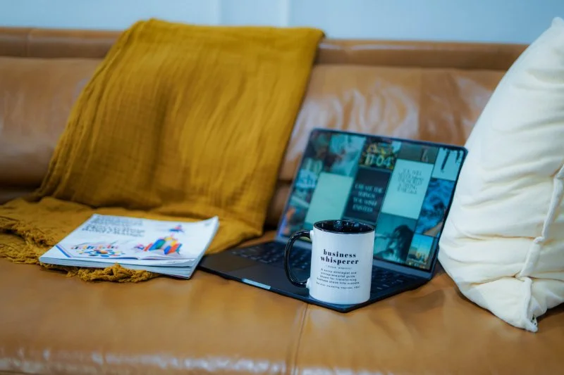 An open laptop, magazine and Business Whisperer mug on a leather couch, the unglamorous setup for figuring out how to write a sales page that converts.