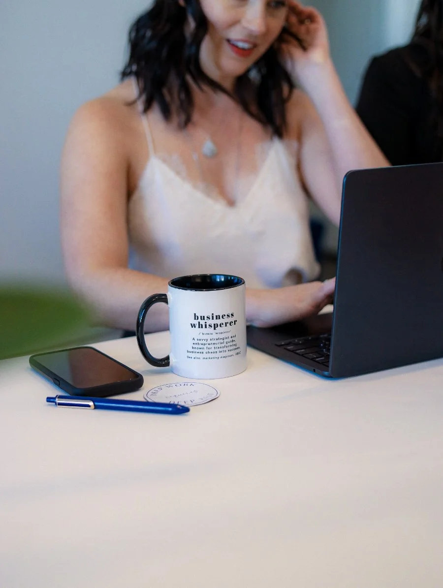 Woman working on a laptop at a clean desk with a branded “business whisperer” mug, phone, and pen nearby. She’s mid-task, slightly smiling, capturing a focused but relaxed work moment that reflects modern, independent business life.