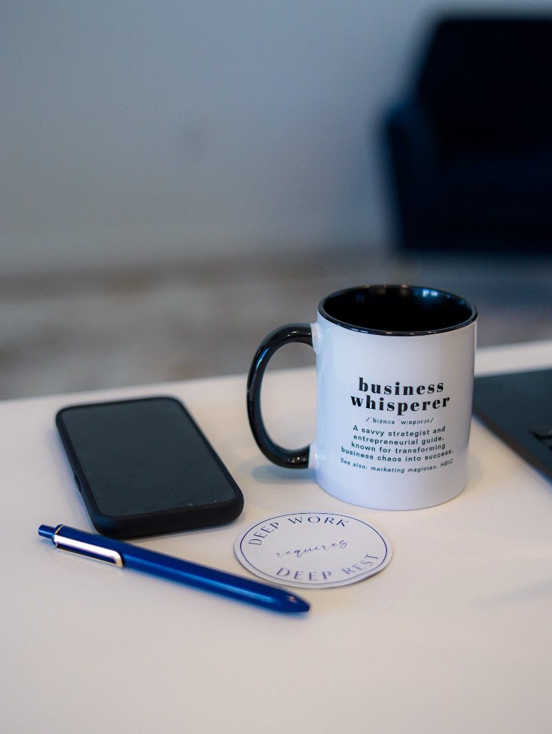 Business Whisperer mug with a dictionary-style definition beside a phone, blue pen, and a "deep work requires deep rest" coaster on a white desk, a calm pause to take before burning it all down and rebuilding your small business messaging.