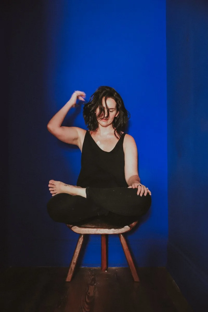 Chelsea Quint sitting cross-legged on a wooden stool against a bold blue backdrop, the calm that comes after you stop forcing your intangible offer into words that don't fit.