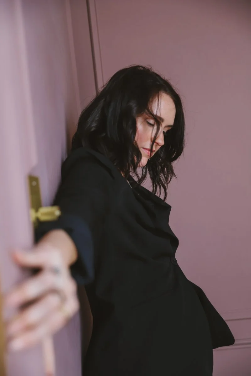 Chelsea Quint, messaging strategist behind The Business Whisperer, in a black blazer against a pink backdrop... helping service providers stop creating more content and start building sales messaging that actually converts.