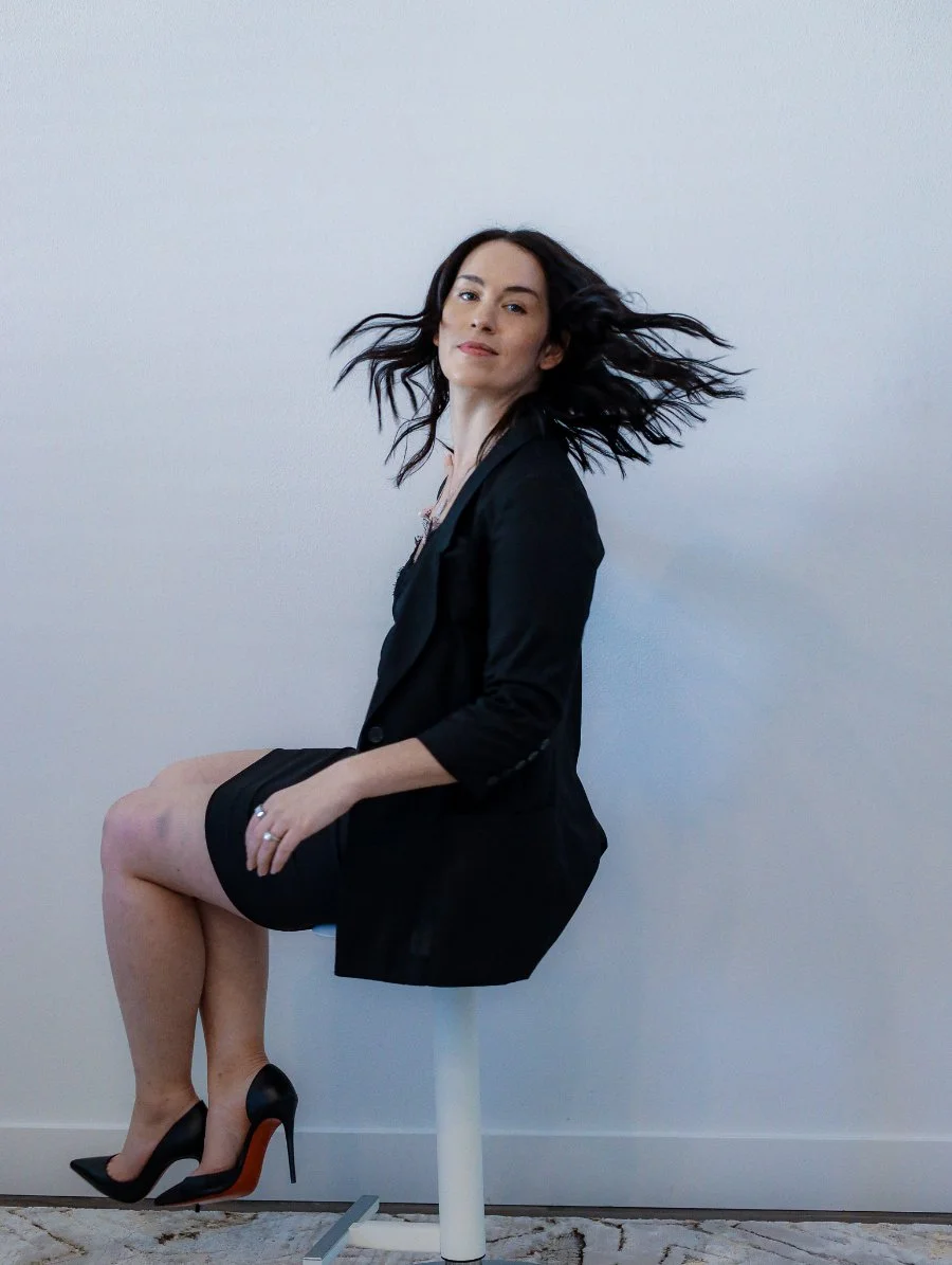 Woman in a black outfit sitting on a stool, hair in motion, looking confident and composed, reflecting clarity and direction after a focused mid-year company audit.