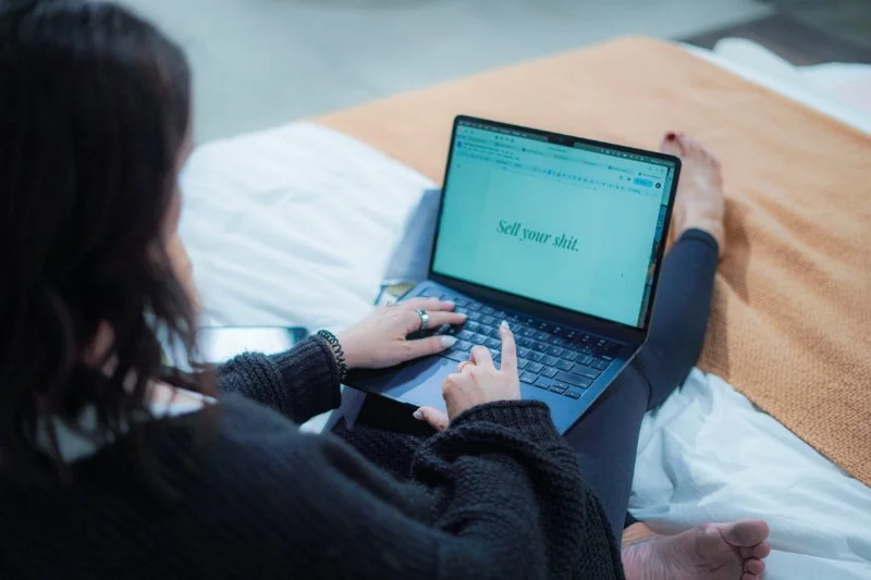 Over-the-shoulder view of a woman sitting cross-legged on a bed, typing on a laptop.