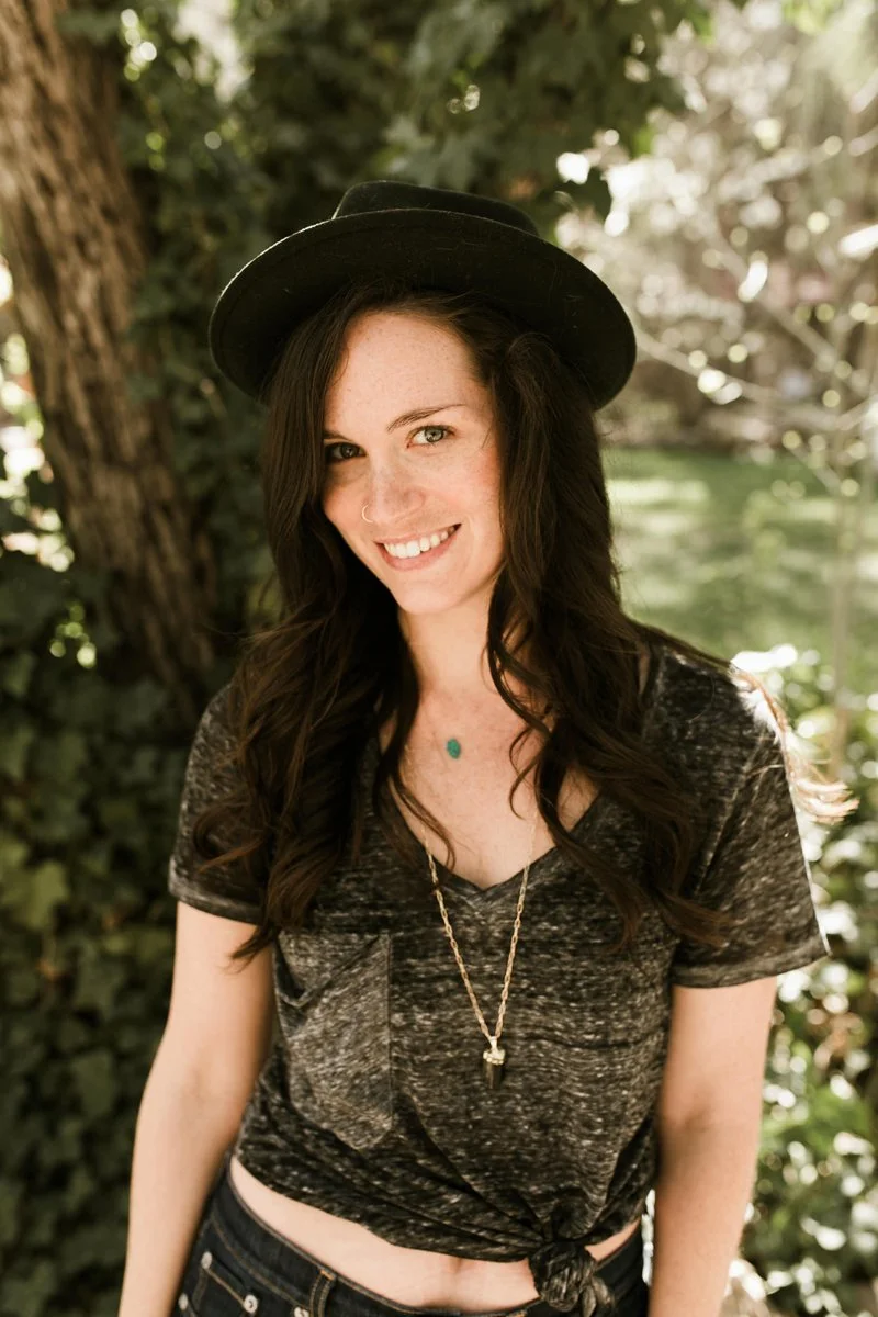 A smiling business owner wearing a black hat stands outdoors beneath leafy trees, creating a warm and approachable brand portrait.