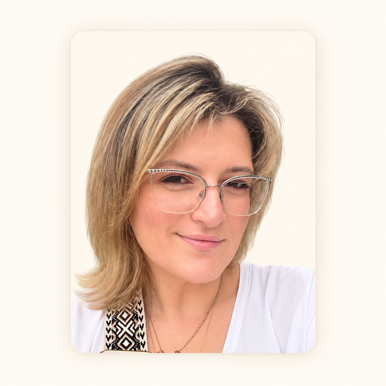 A woman with blonde hair wearing glasses and a white top, smiling against a plain light background.