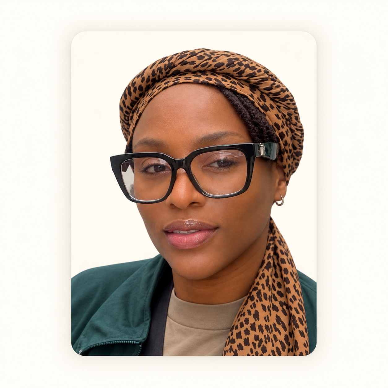A woman with a leopard print headscarf, large black glasses, and a small hoop earring, wearing a green jacket and beige shirt, posing against a plain background.
