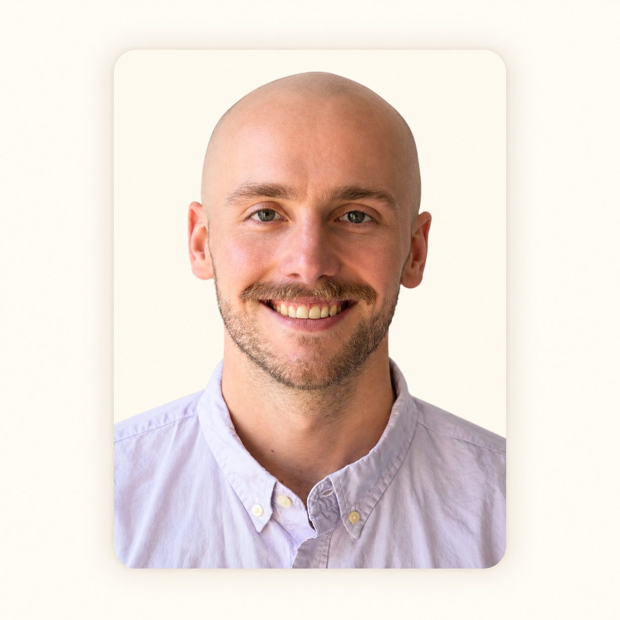 A smiling young man with a shaved head, light beard, wearing a light purple button-up shirt against a plain white background.