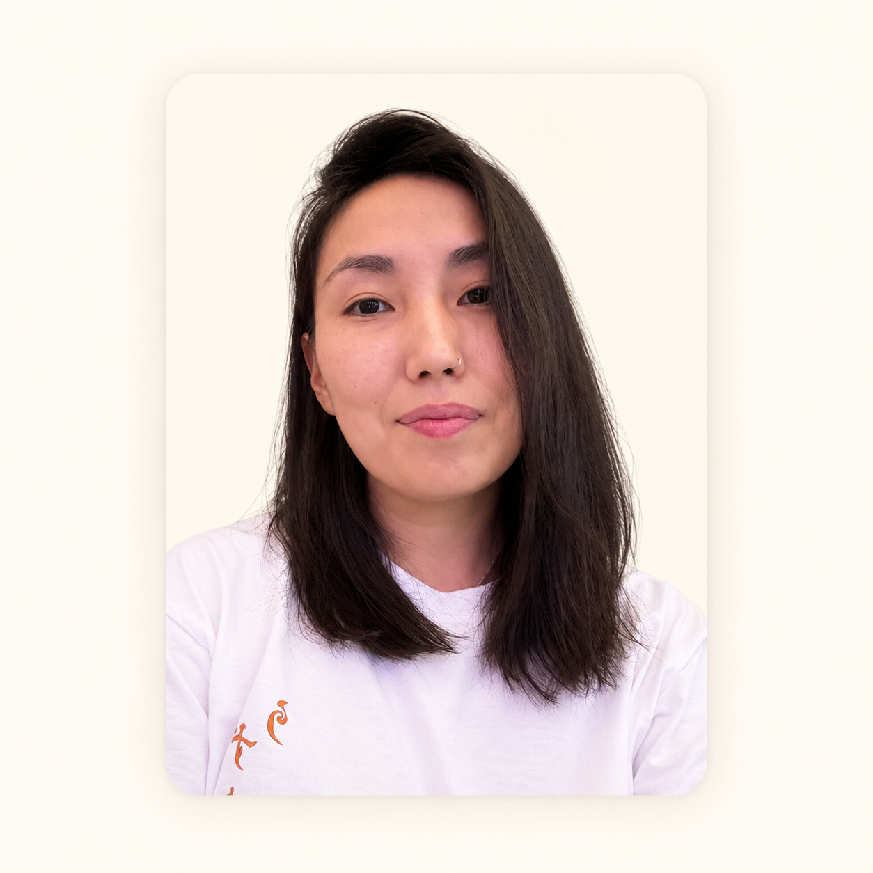Close-up of a young woman with dark brown, shoulder-length hair, wearing a white shirt with orange text, standing against a plain white background.