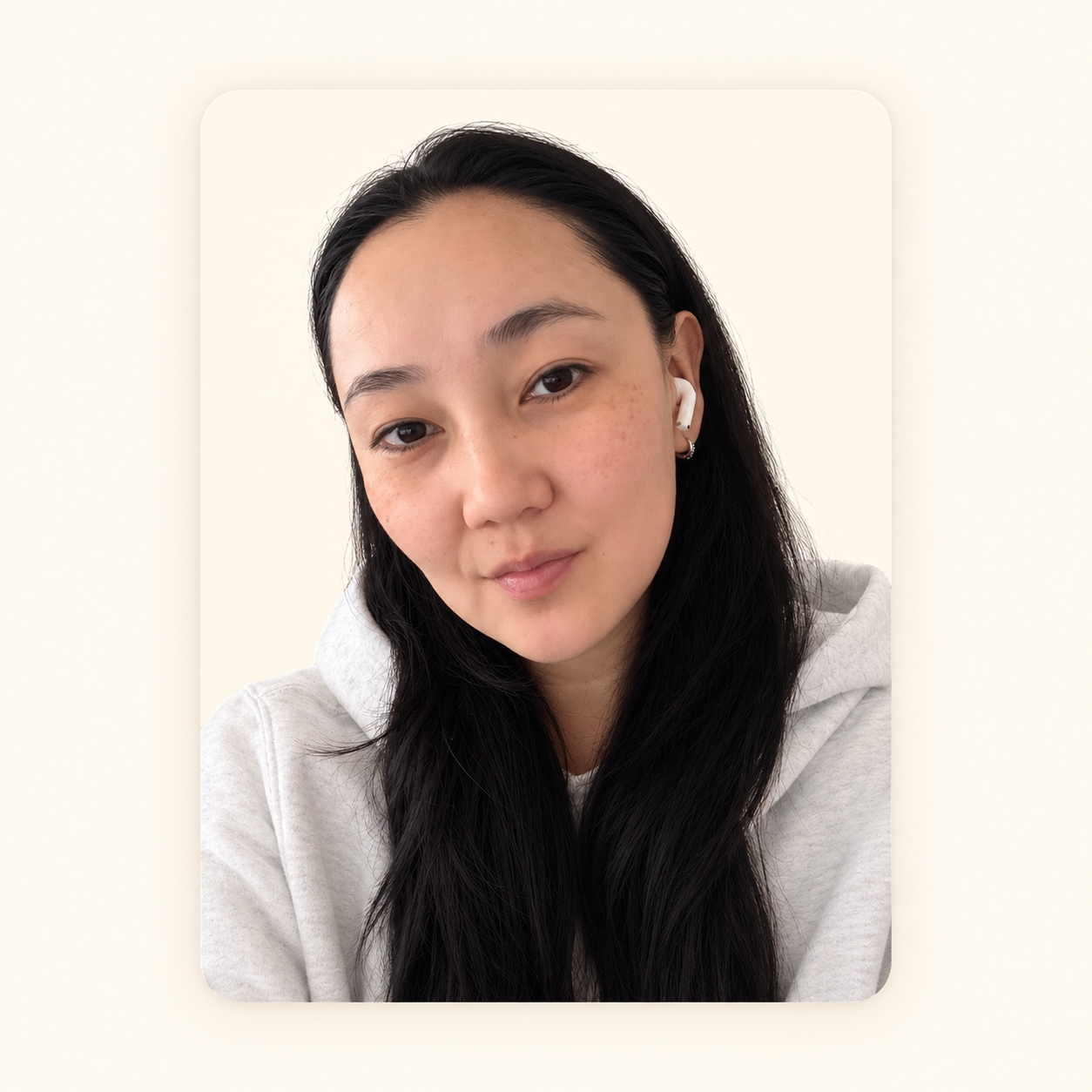 A young woman with long dark hair, wearing a gray hoodie and AirPods, looking at the camera with a neutral expression against a plain light background.