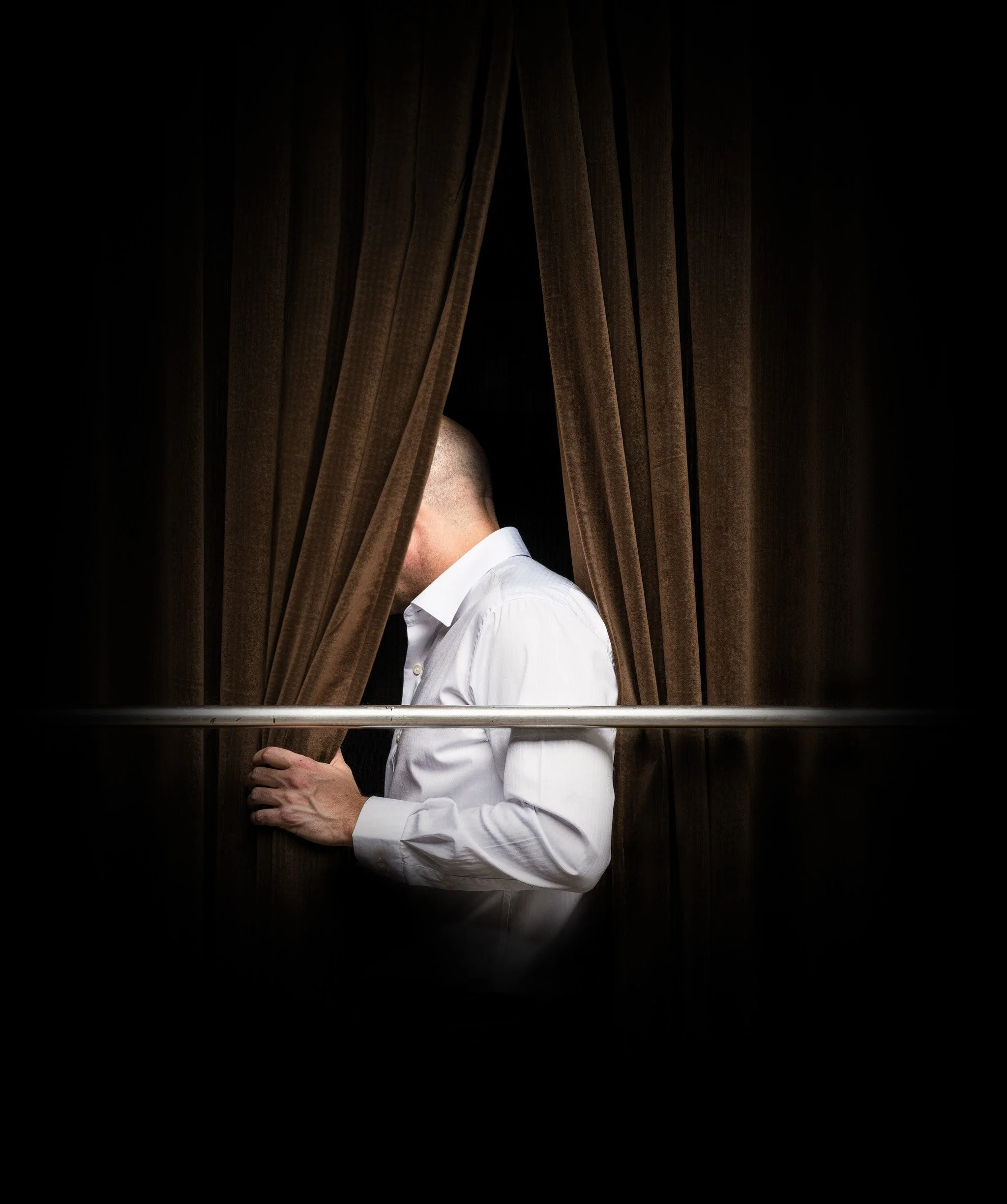 A person with a shaved head and light skin wearing a white shirt, partially visible behind brown curtains, with only part of their head and arm showing.