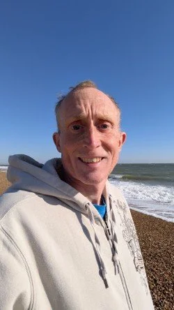Person in a hoodie smiling on a pebble beach with blue sky and ocean waves in the background.