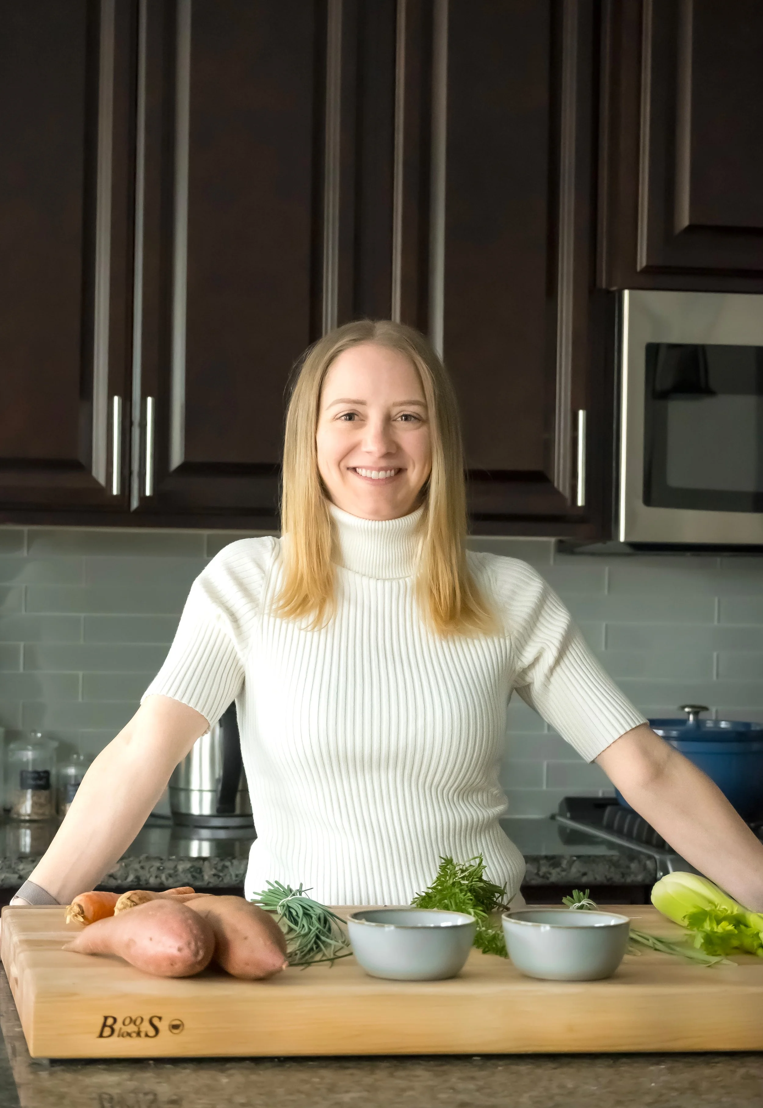A woman smiling in a kitchen, standing behind a wooden cutting board with fresh vegetables and herbs, including sweet potatoes, green onions, and rosemary.