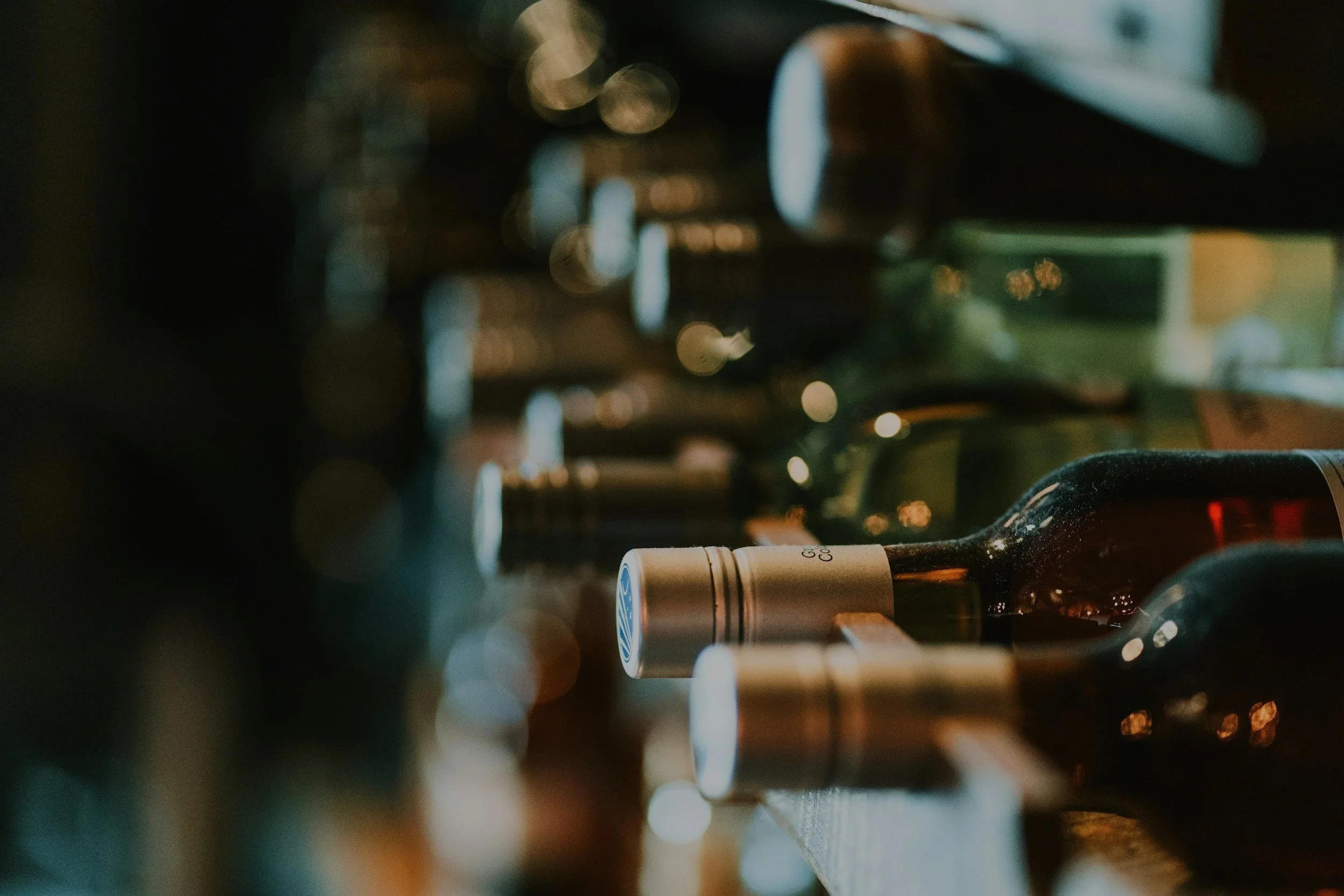 A row of wine bottles lying on their sides on a wine rack, with one bottle near the front in focus, featuring a dark glass body and a silver capsule around the neck.