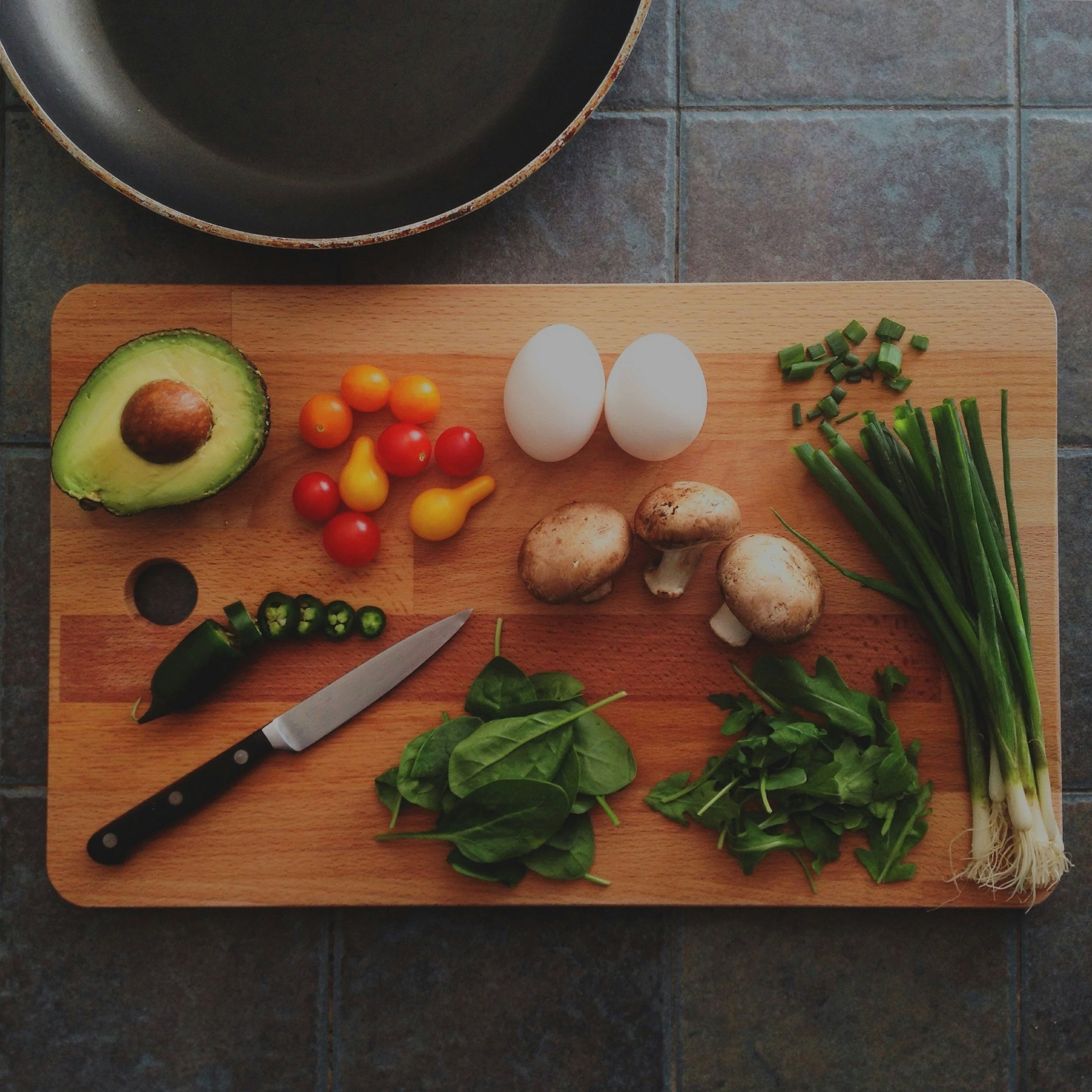 Cutting board with avocado, cherry tomatoes, eggs, mushrooms, green onions, jalapeno, spinach, arugula, and a knife, on a tiled kitchen countertop.