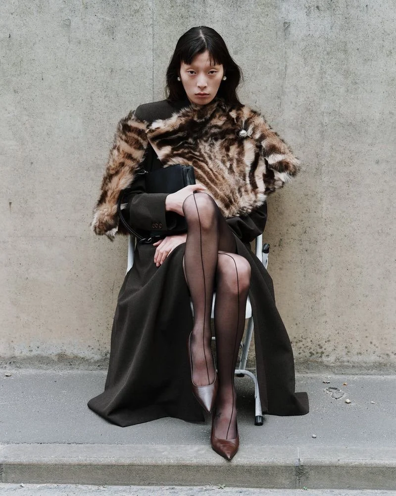 Femme assise contre un mur en béton, portant un manteau en fourrure et une robe longue noire avec des collants transparents et des talons hauts marron