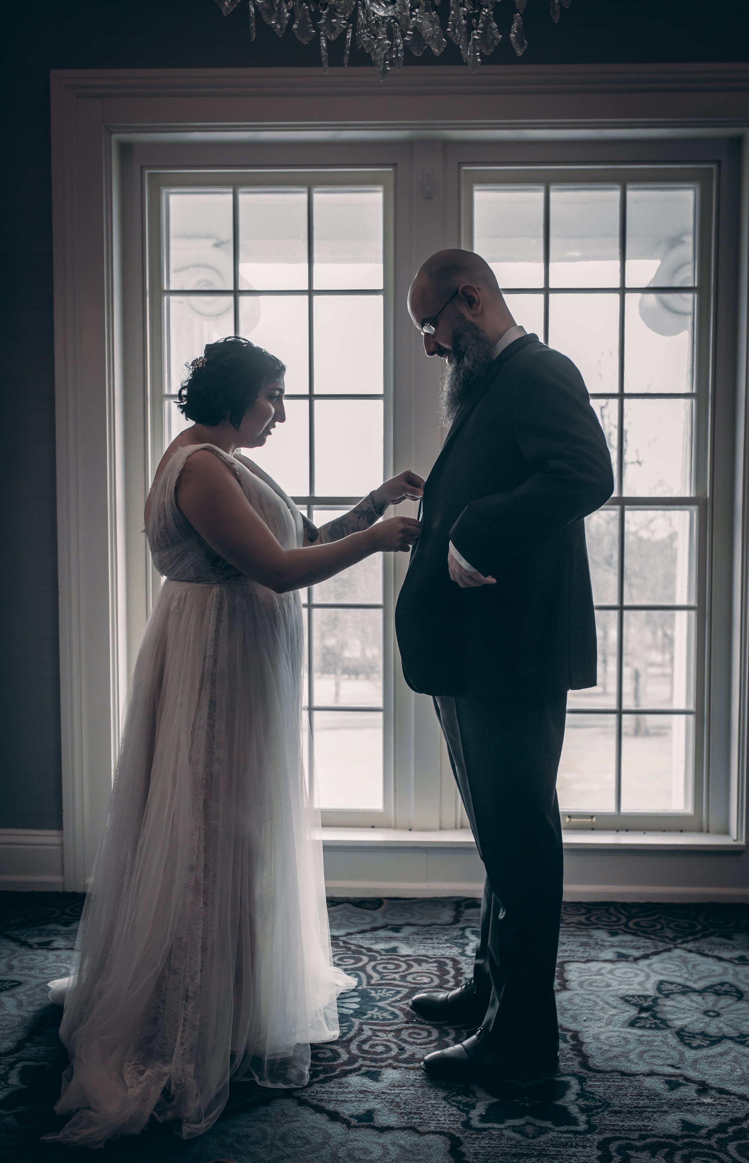 The bride adjusts her groom's suit jacket as they stand together in beautiful window light before their ceremony.