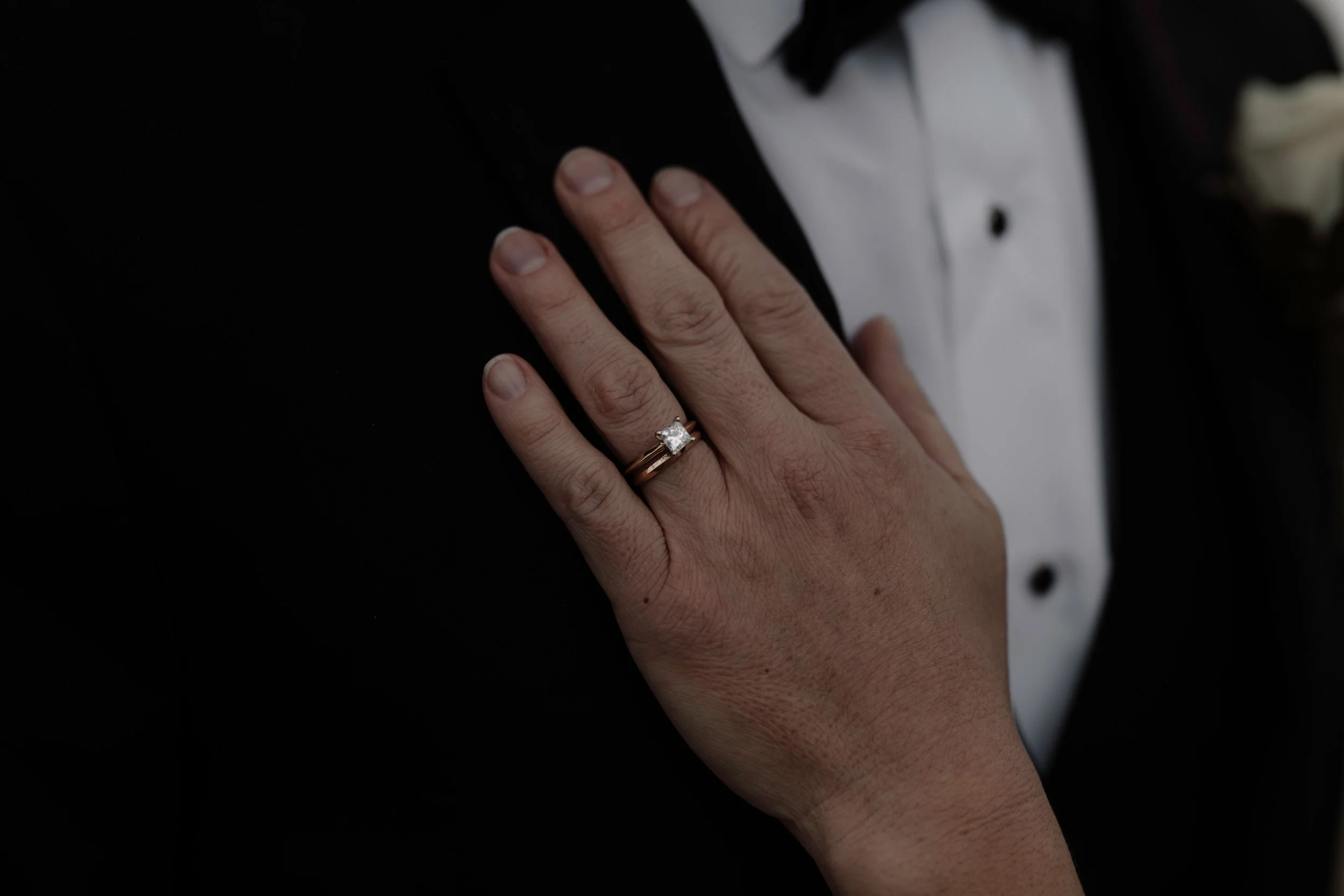 Elegant wedding detail photography featuring a diamond engagement ring on a bride's hand resting on a groom's tuxedo.