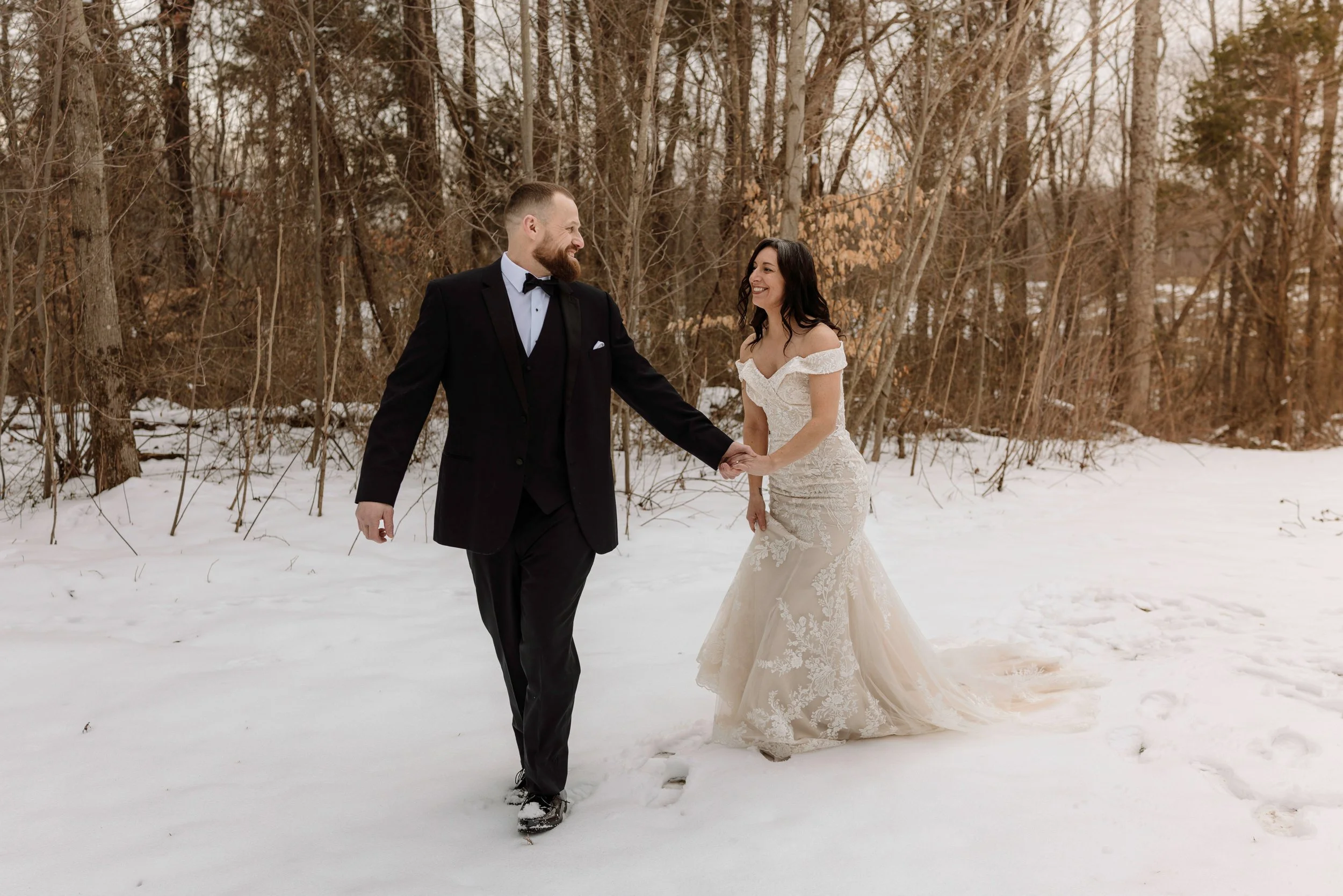 "Bride in a lace wedding dress and groom in a tuxedo walking through a picturesque snowy landscape on their wedding day.