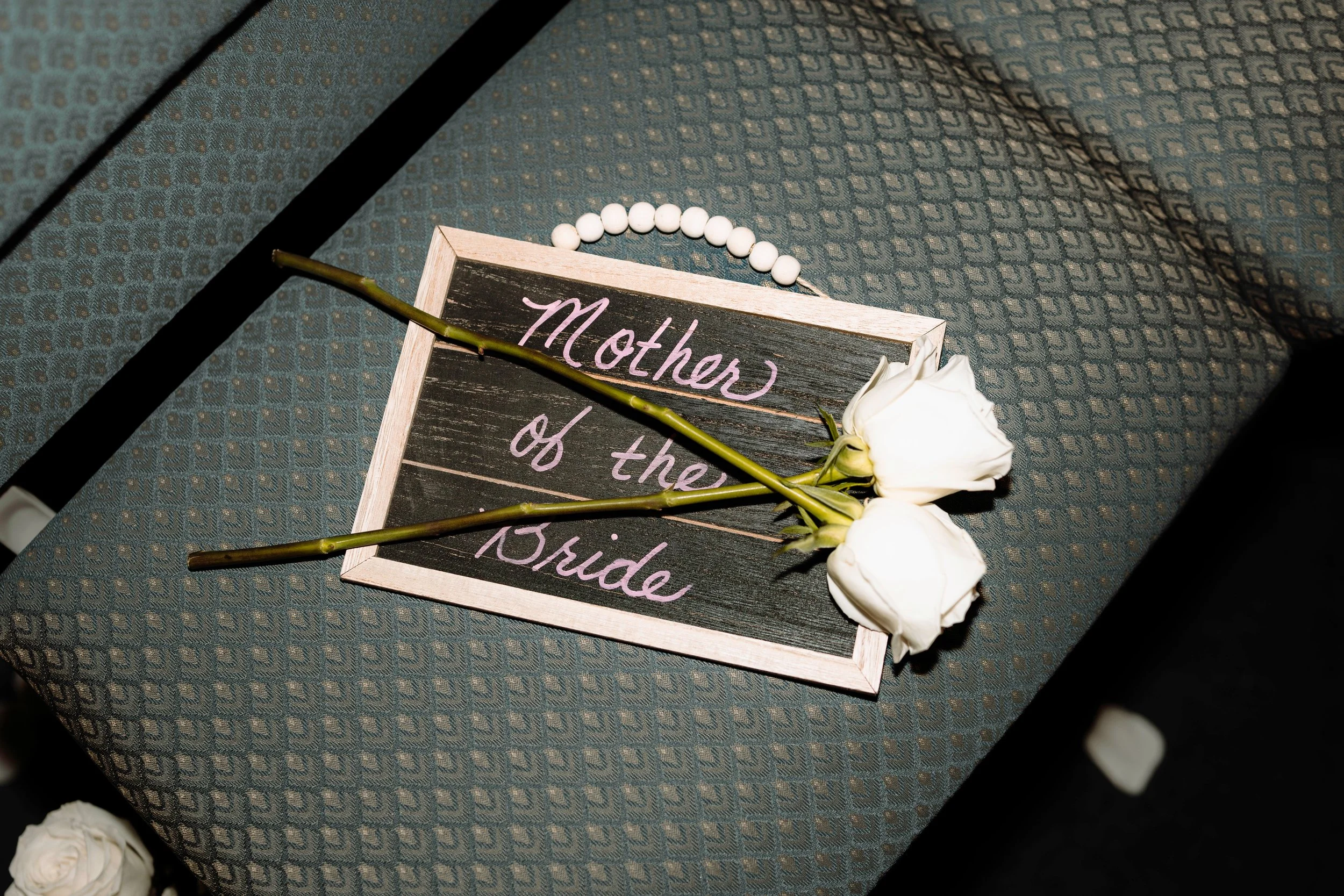 Wedding detail photography of a 'Mother of the Bride' chalkboard sign adorned with two white roses and pearl beads.