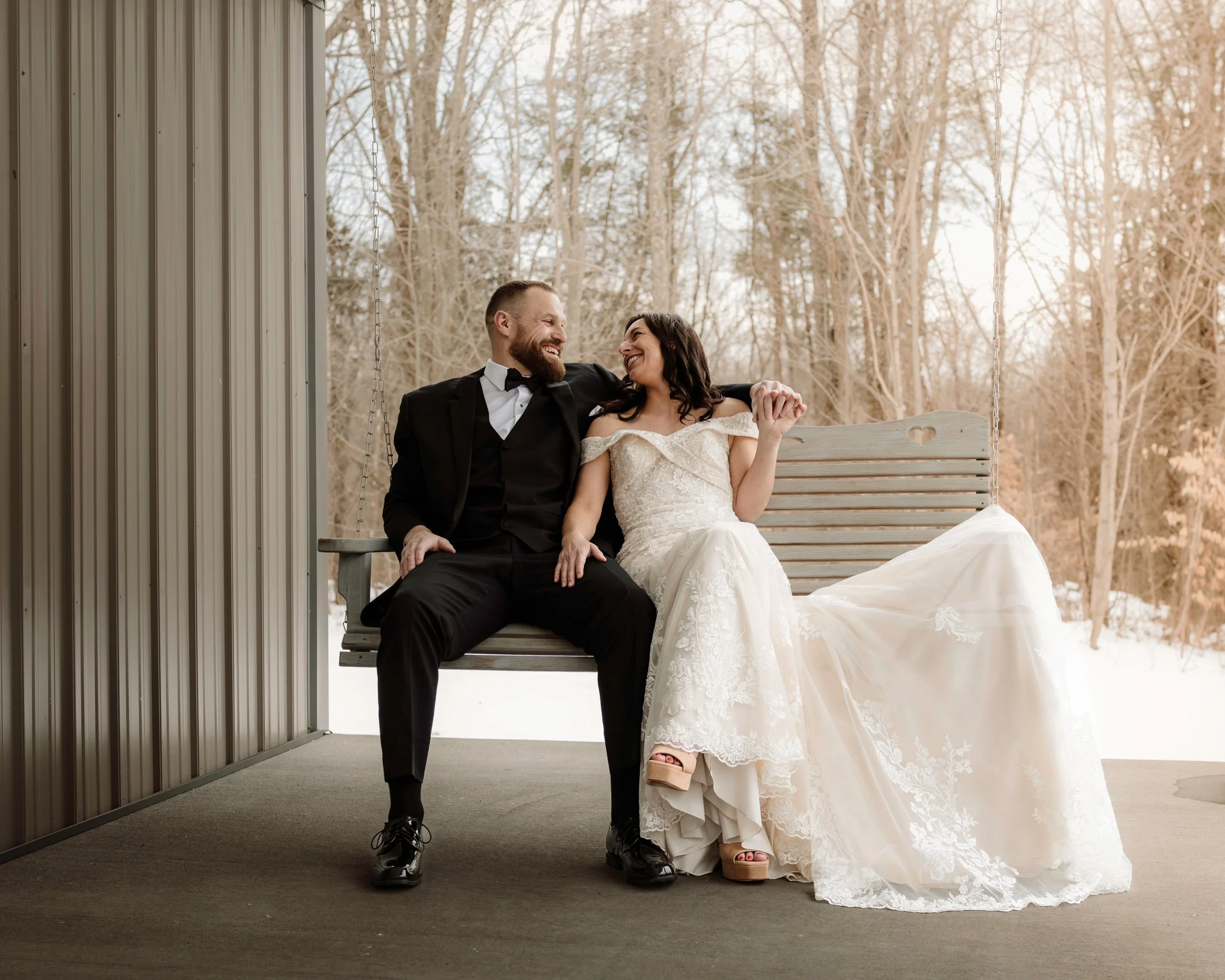 Romantic wedding portrait of a bride and groom sharing a laugh on a swing bench with a beautiful snowy forest background.