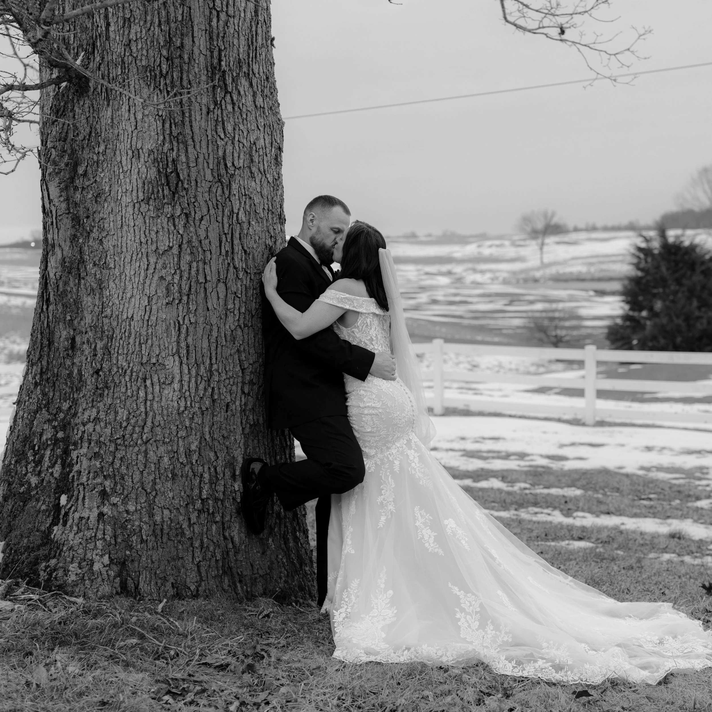Romantic black and white wedding photo capturing a couple kissing while leaning against a tree trunk in a beautiful snowy landscape.