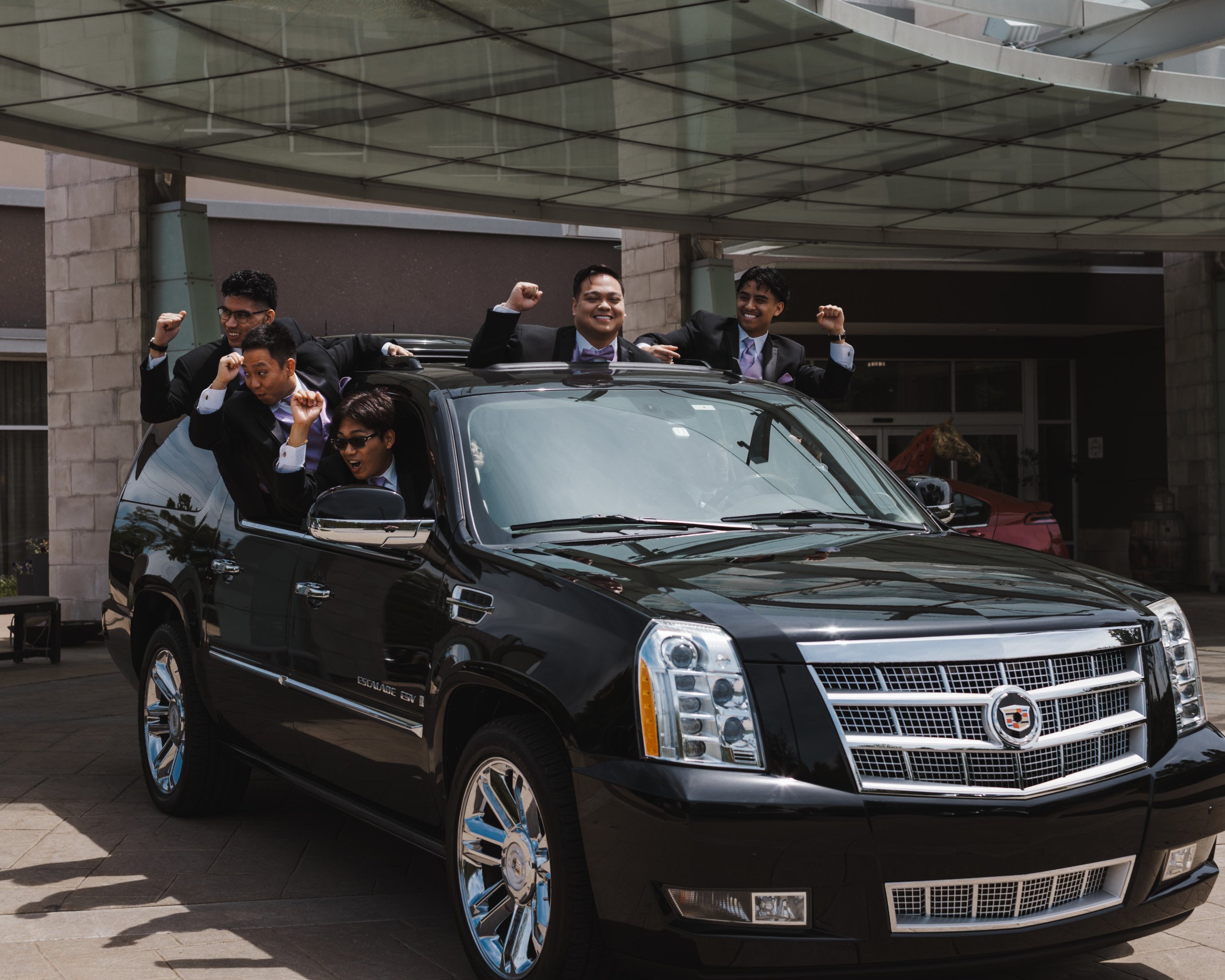 Still from a wedding film, showing groomsmen celebrating exuberantly from a black Cadillac Escalade SUV.