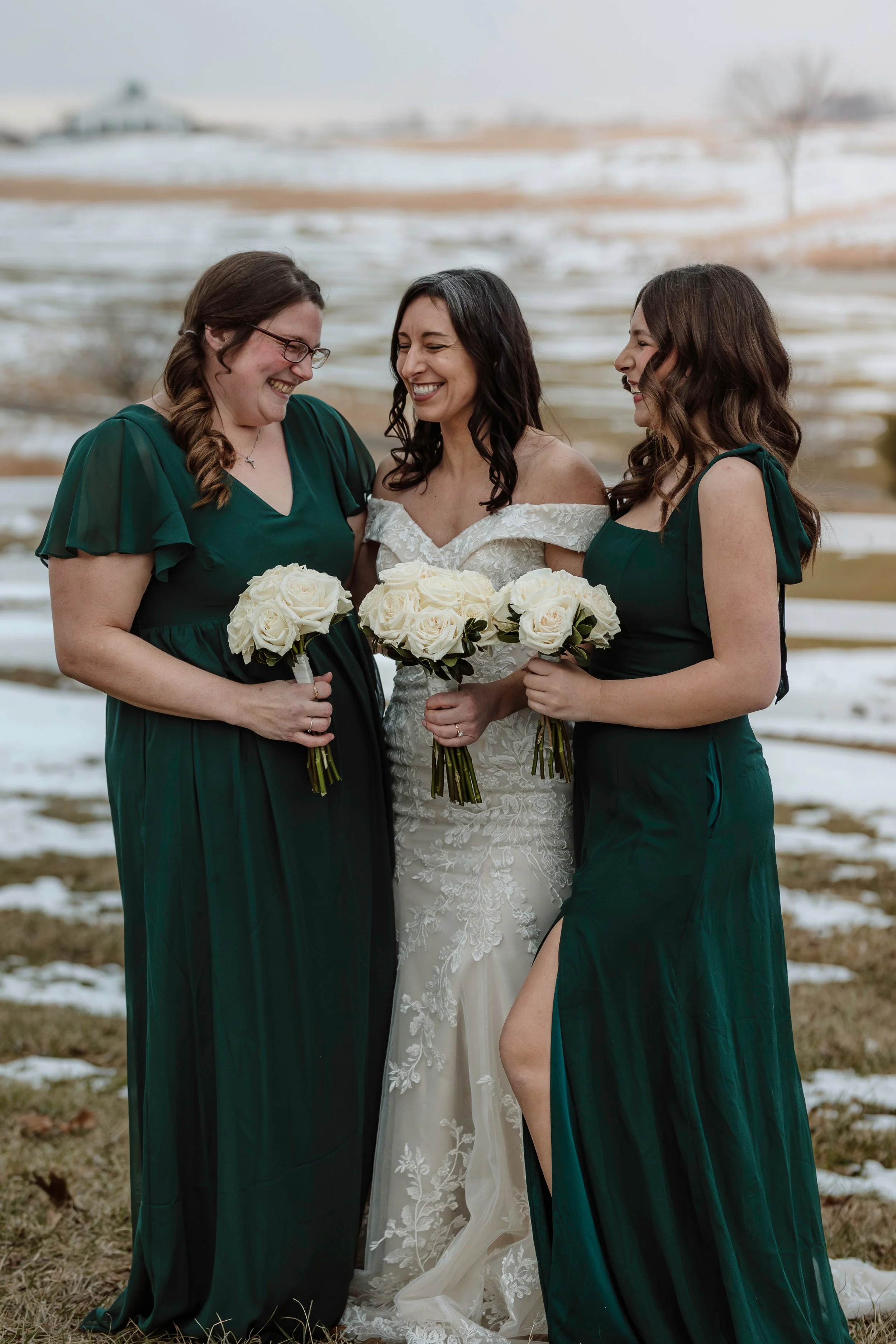 Bride smiling with her bridesmaids in long dark green dresses, holding white rose bouquets in a snowy setting.