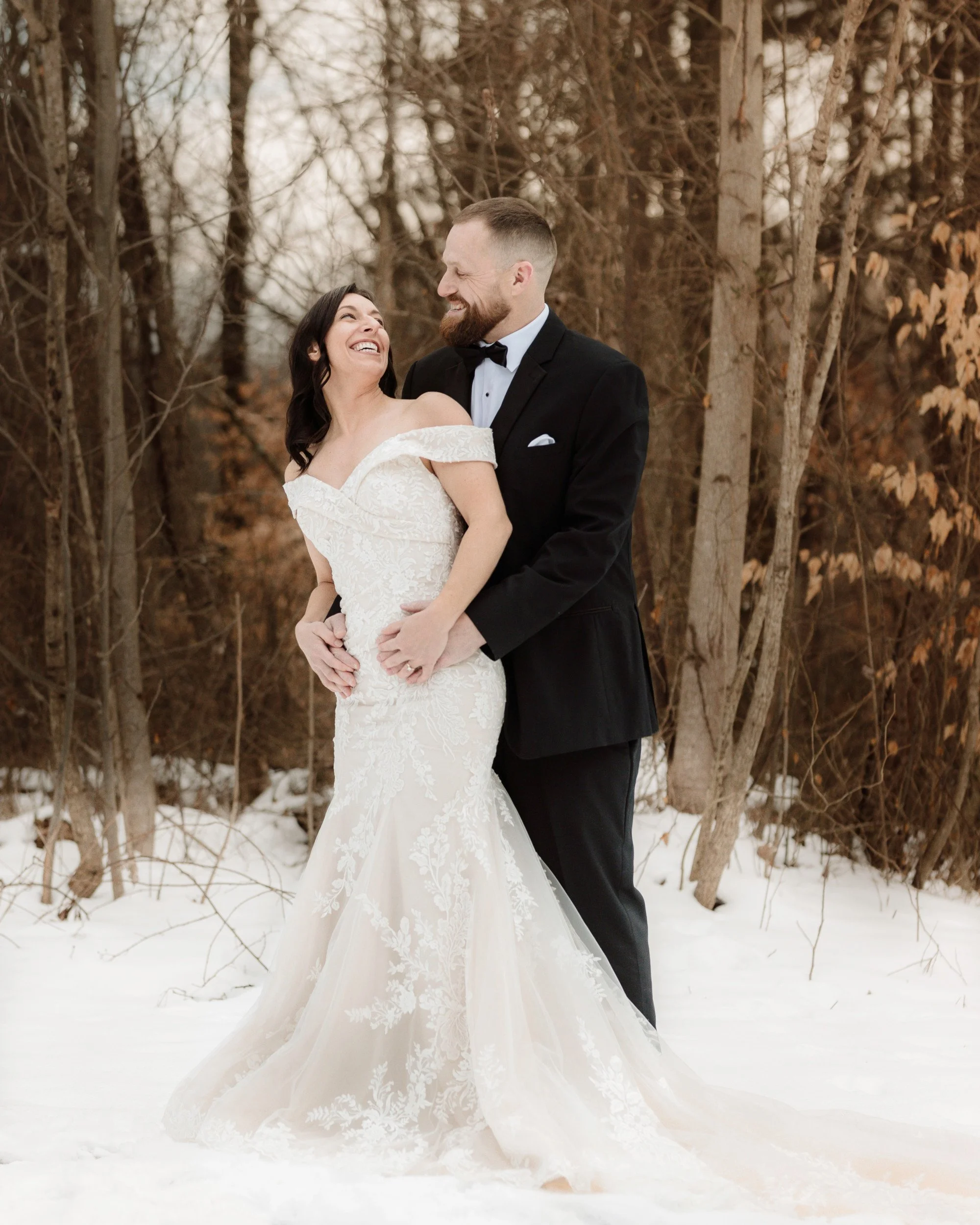 Bride in a white lace wedding dress and groom in a tuxedo, posing for a portrait in a serene snowy forest.