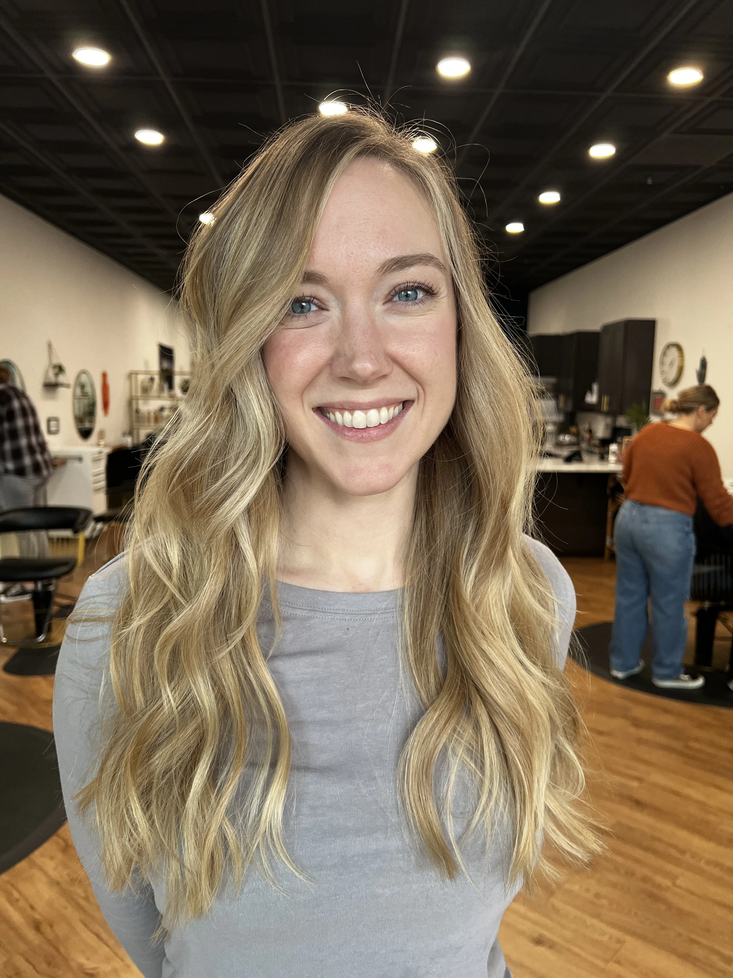 A person with long, wavy blond hair smiling in a salon setting with wooden flooring and modern decor.