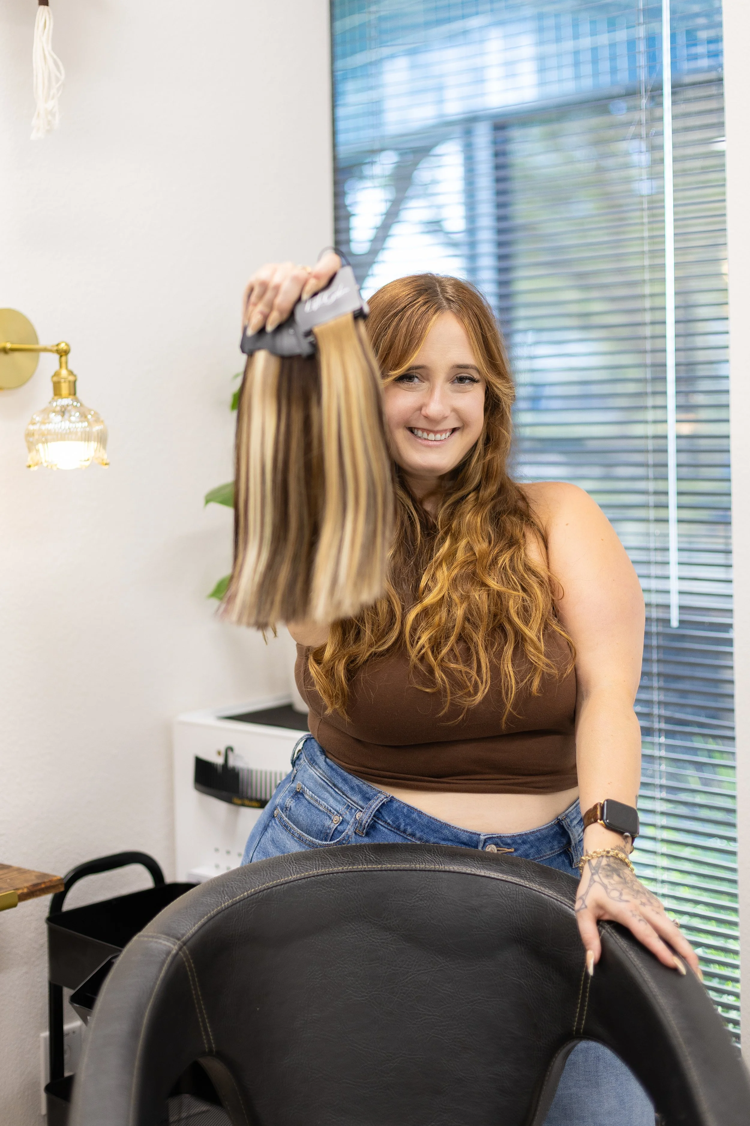 Hair extensions in various colors and textures hanging on a rack above a wicker chair and surrounded by potted plants.