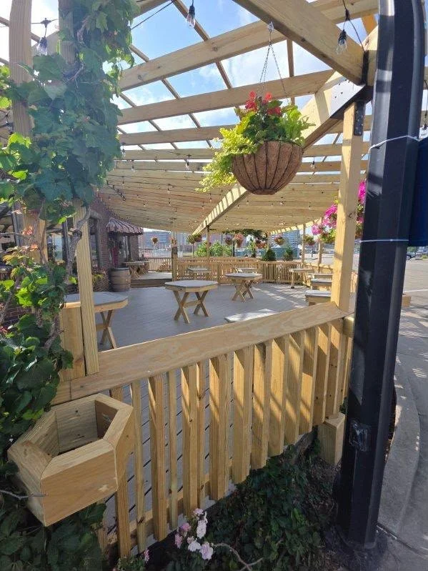 A wooden outdoor patio with tables, benches, and hanging flower baskets, partially covered with a wooden pergola. There are plants and flowers around the patio and a sidewalk with a street visible in the background.