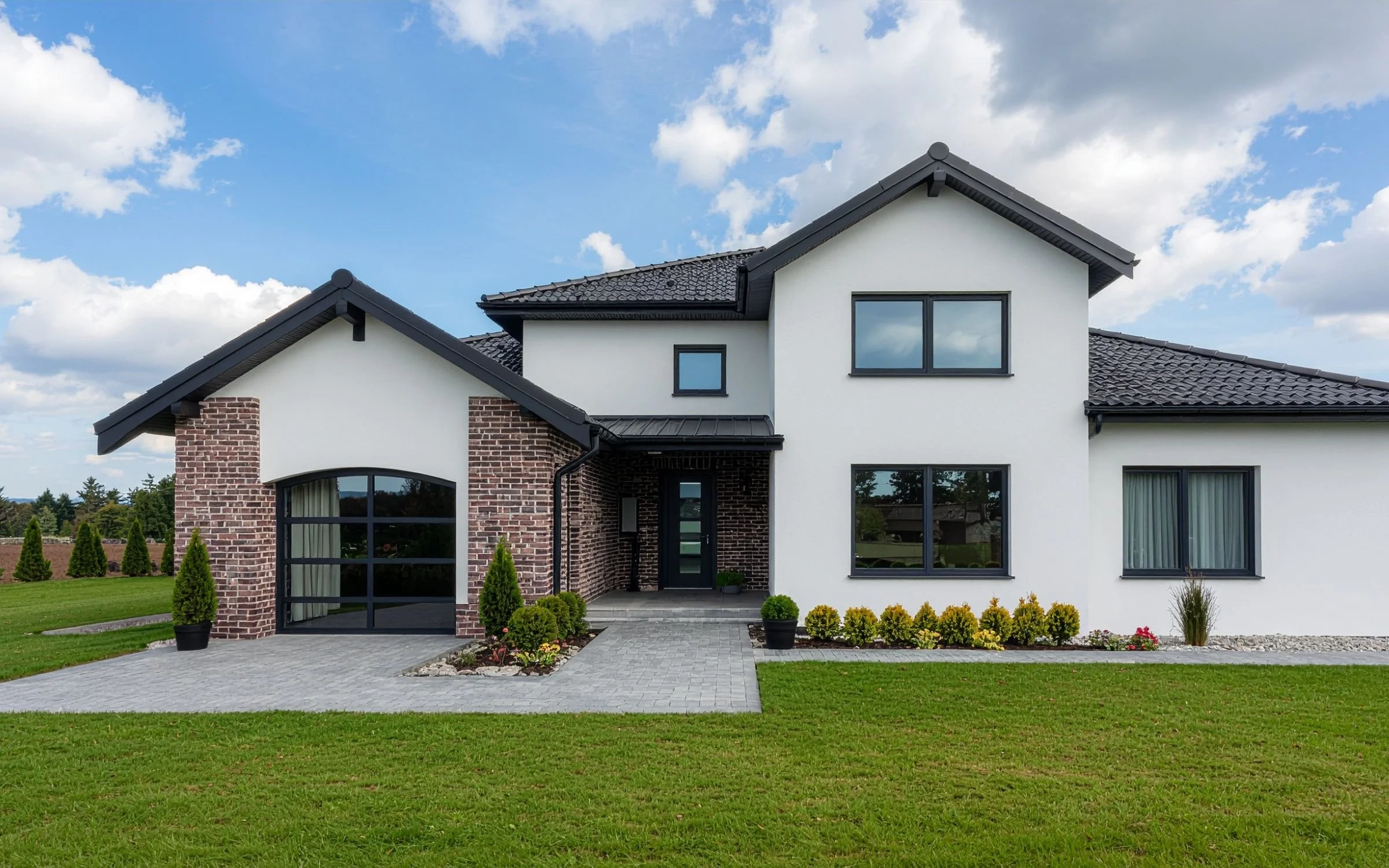A modern two-story house with white and brick exterior, black-framed windows, and a black tile roof, surrounded by a well-manicured lawn and small bushes under a blue sky with scattered clouds.