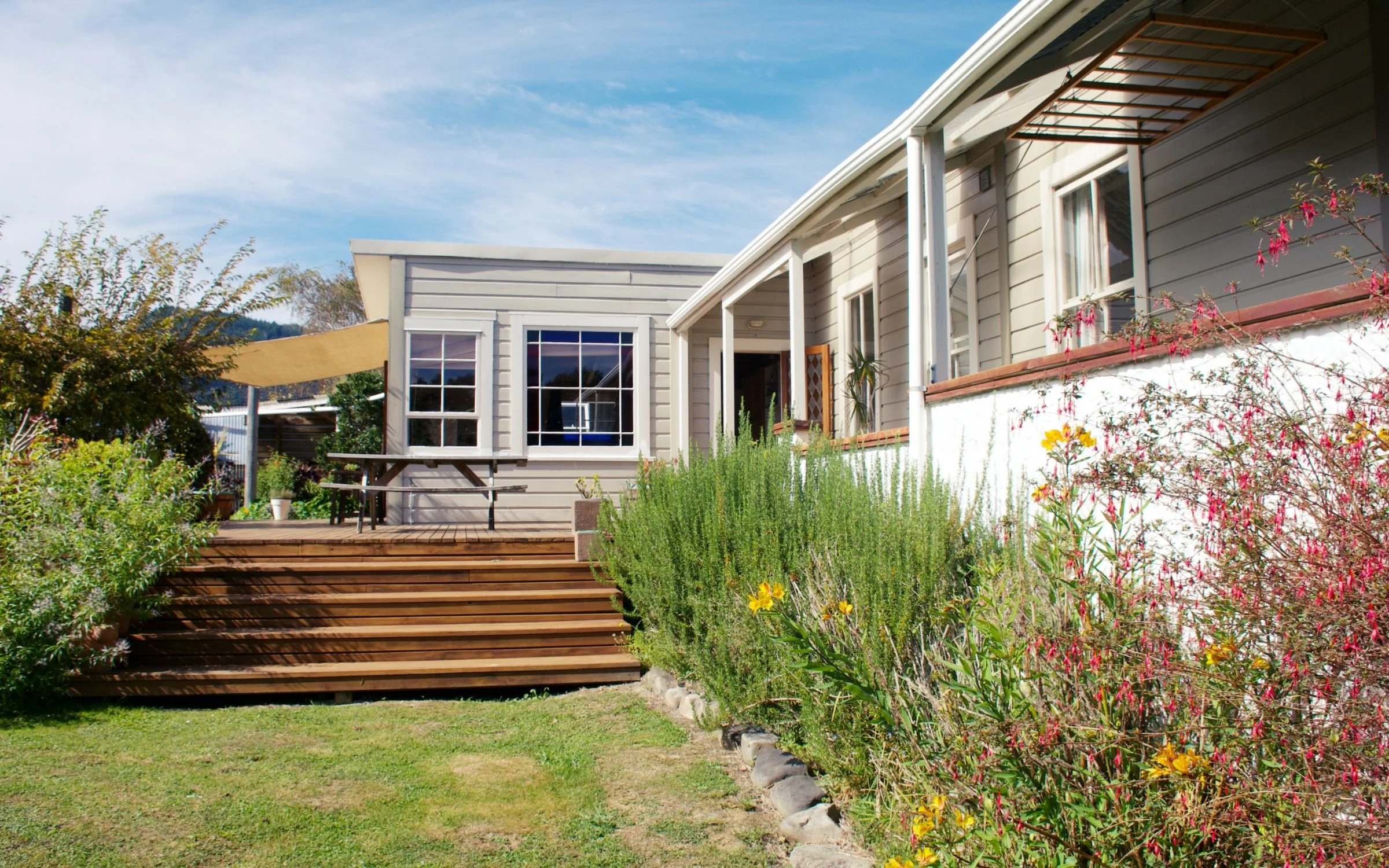 Backyard garden with wooden stairs leading to a house with gray siding, white trim, and a patio with plants and flowers, clear blue sky.