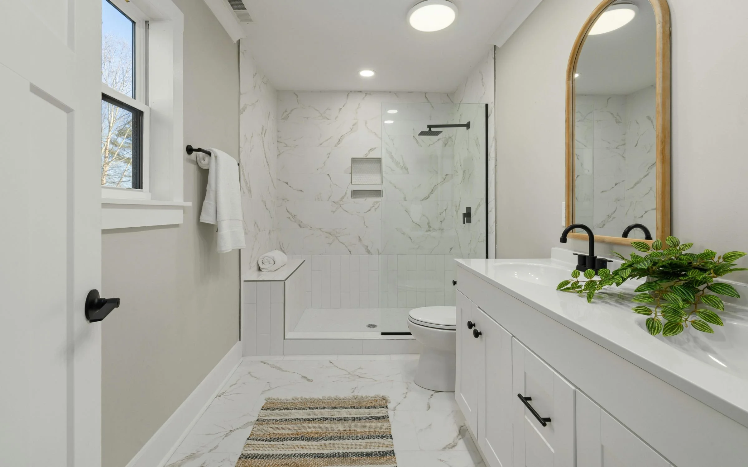 Modern bathroom with white marble walls and flooring, black fixtures, a large mirror with a wooden frame, a white vanity with a black faucet, a potted plant, and a walk-in shower with a black showerhead.
