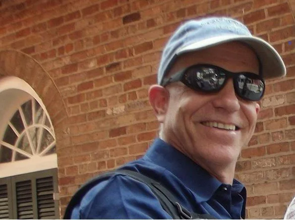 Smiling man in sunglasses and a light baseball cap, wearing a blue jacket, standing outside in front of a brick building with a window.