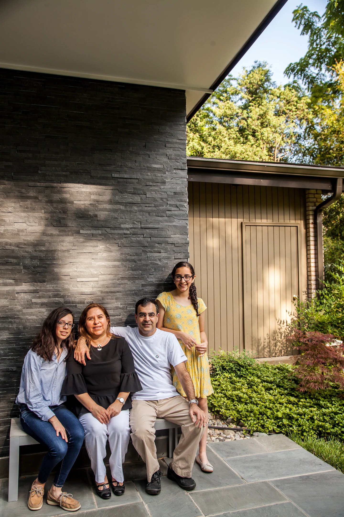 Group of four people sitting and standing outside in front of a modern house with black stone wall and garden.