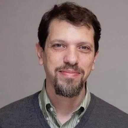 Headshot of a man with brown hair and a goatee, wearing a striped shirt and a gray sweater, smiling against a plain background.