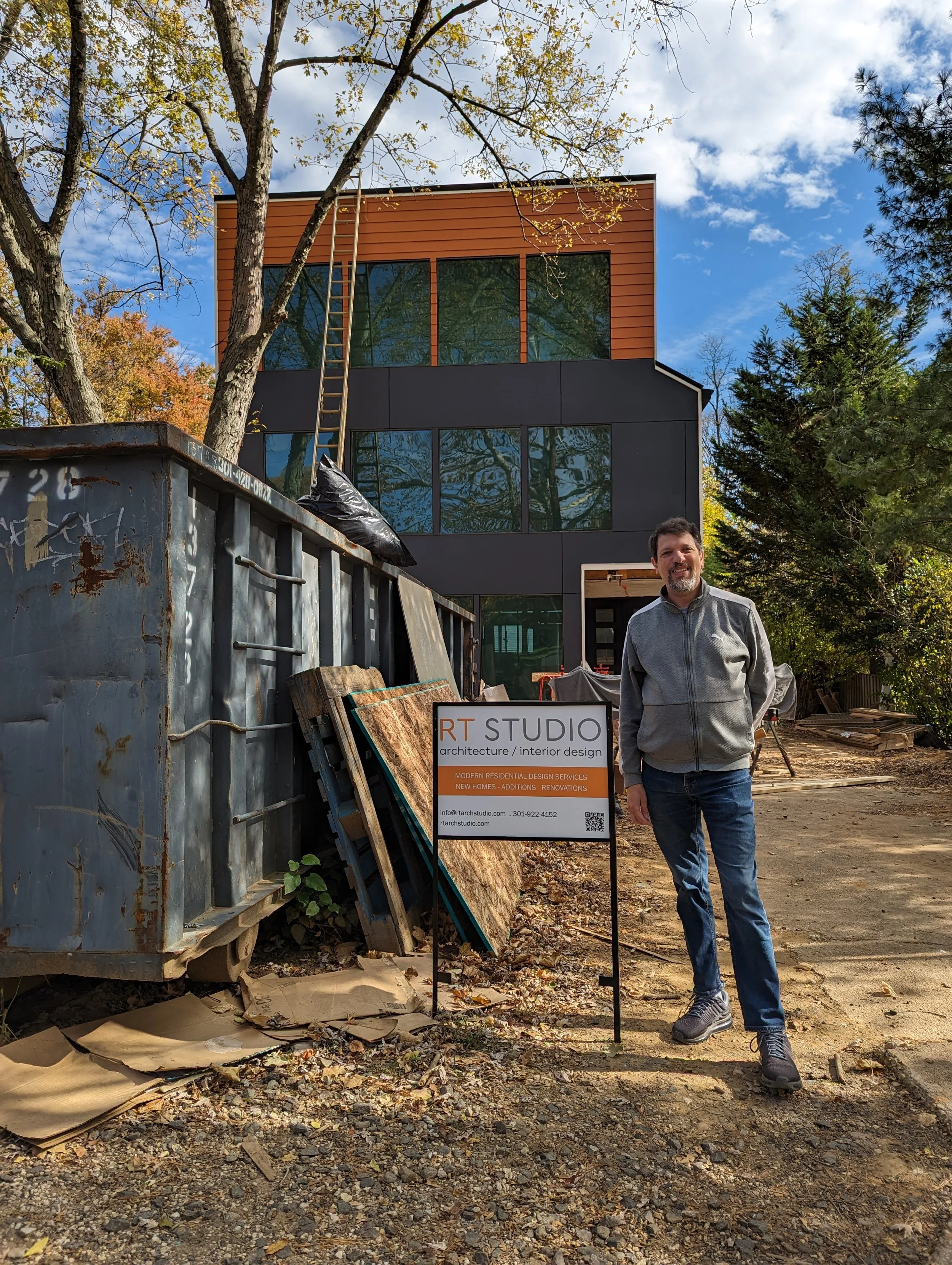 A man stands on a construction site in front of a sign that reads 'RT Studio architecture / interior design'. In the background, there's a modern house with large windows, surrounded by trees with fall foliage, and a construction dumpster and materials nearby.
