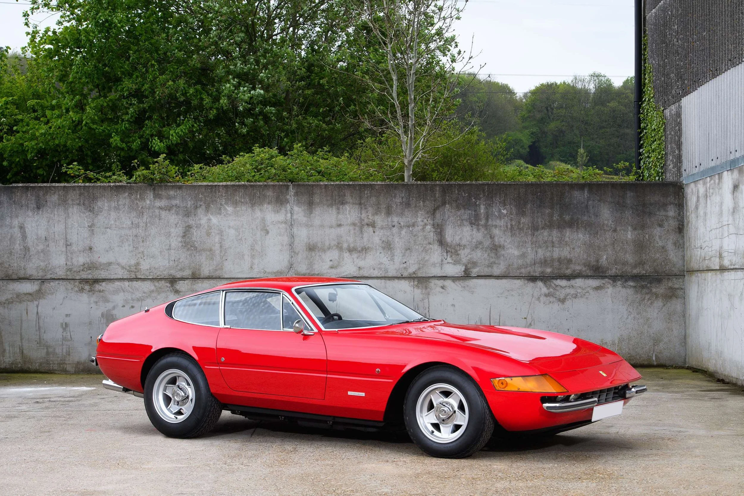 A classic red sports car parked outdoors near a concrete wall, with green trees and a cloudy sky in the background.