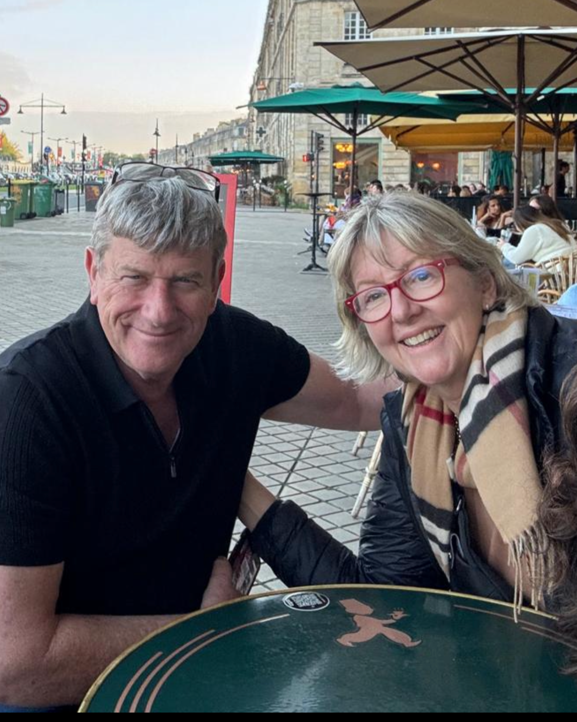 A smiling man and woman sitting at an outdoor cafe table on a city street, with people dining and umbrellas in the background.