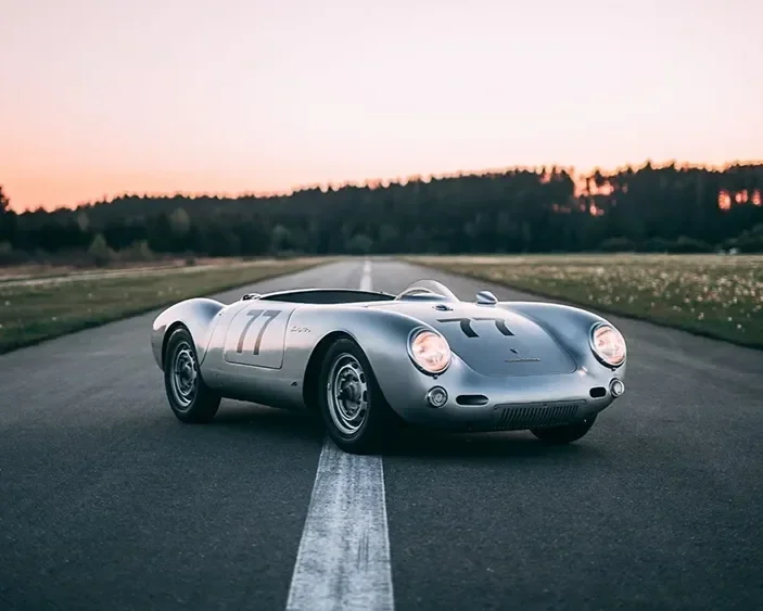 A classic silver race car with the number 77 on its side, parked on an empty road at sunset, with trees and a pink sky in the background.