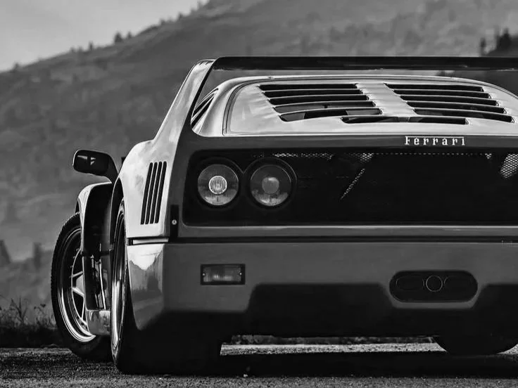 Black and white photo of the rear of a classic Ferrari sports car on a road with a mountainous background.