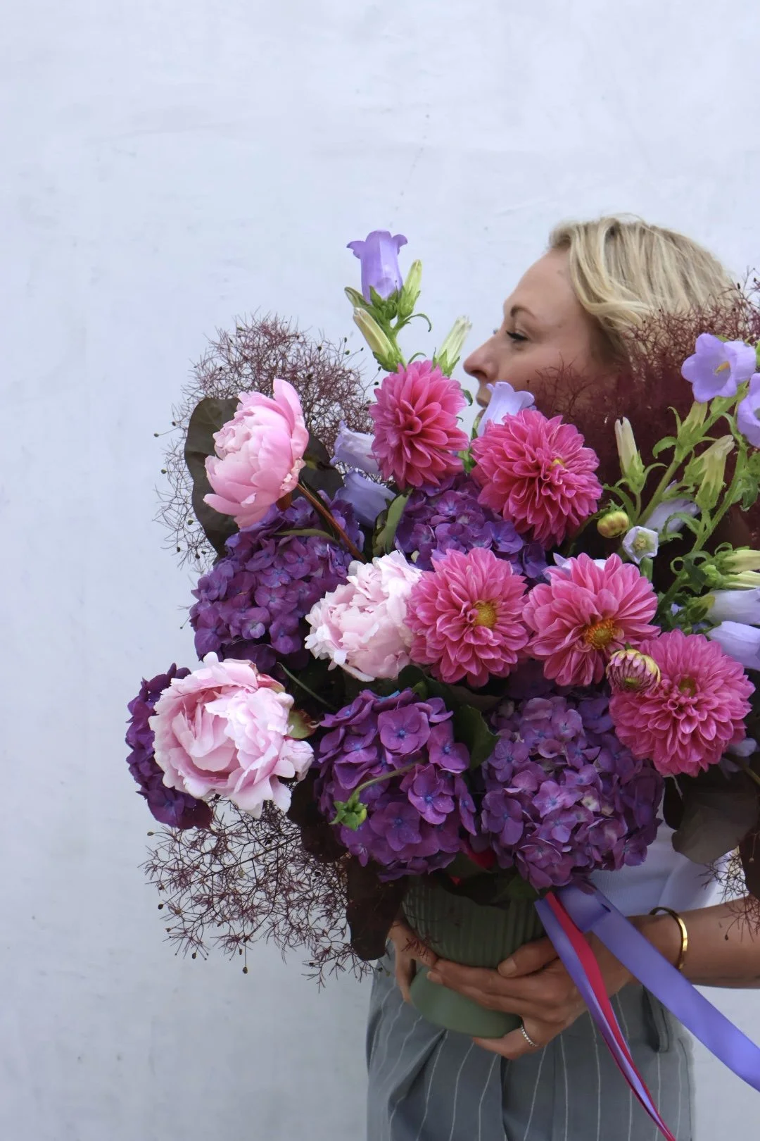 A woman holding a large bouquet of pink, purple, and lavender flowers against a plain, light background.