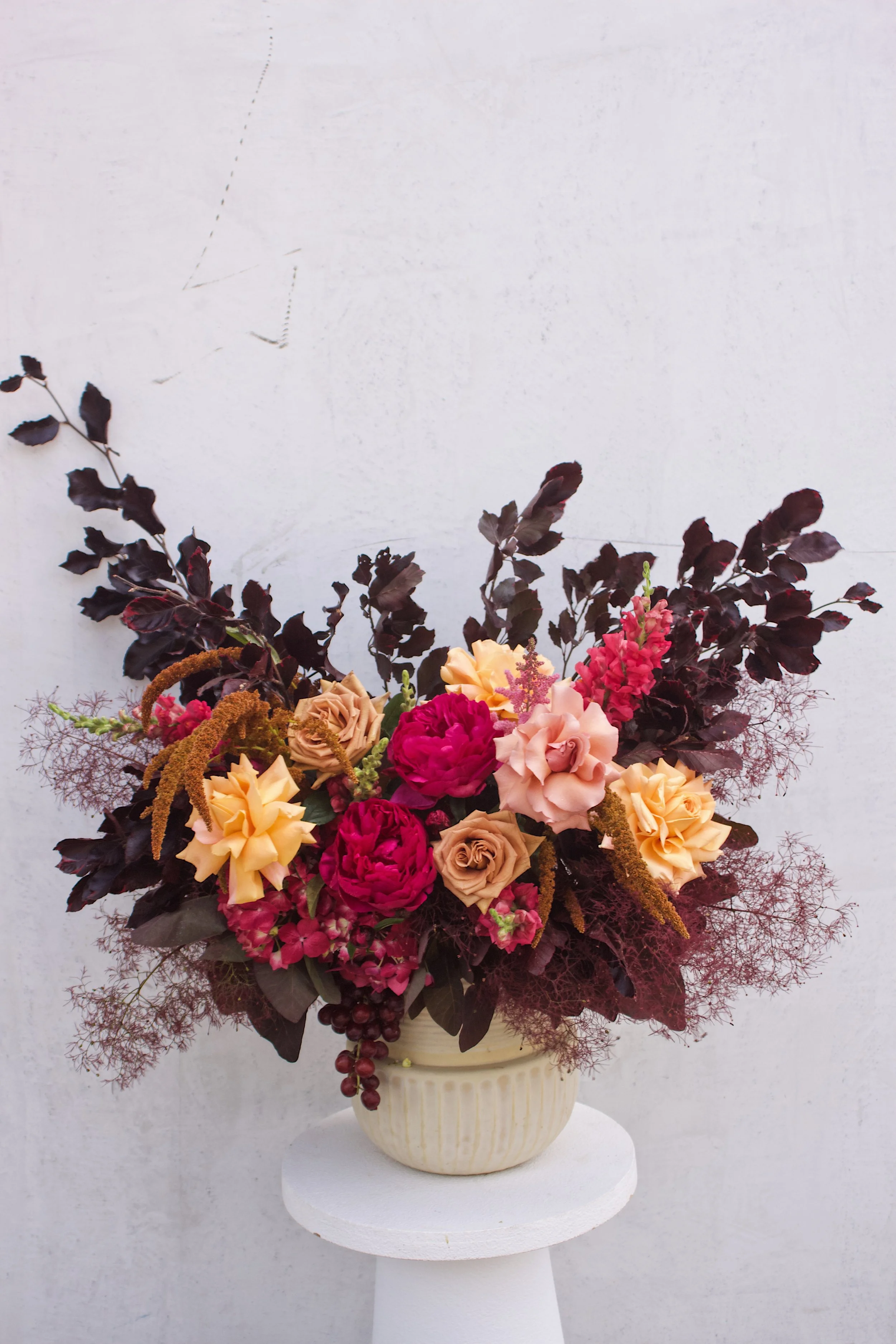A floral arrangement in a beige ceramic vase featuring dark purple leaves, pink and peach roses, and pink peonies, set against a plain light background.