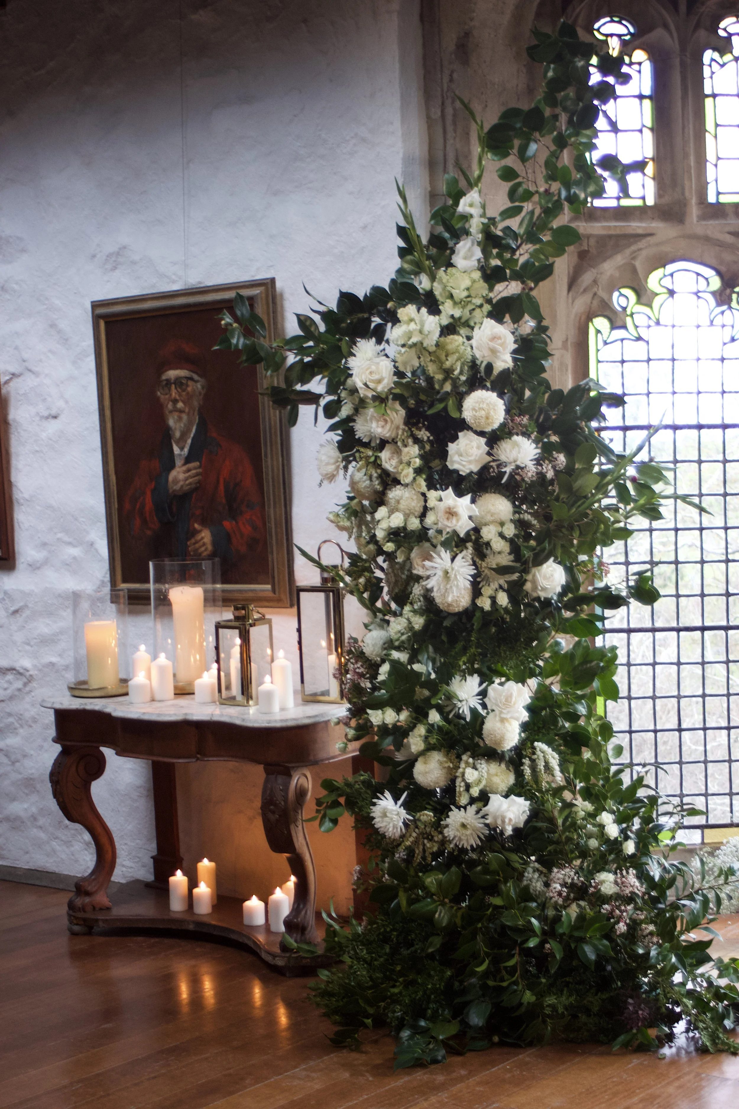 Indoor scene with a large white floral arrangement, lit candles, and a portrait of an older man wearing a red robe and glasses on the wall behind. Sunlight filters through stained glass windows.