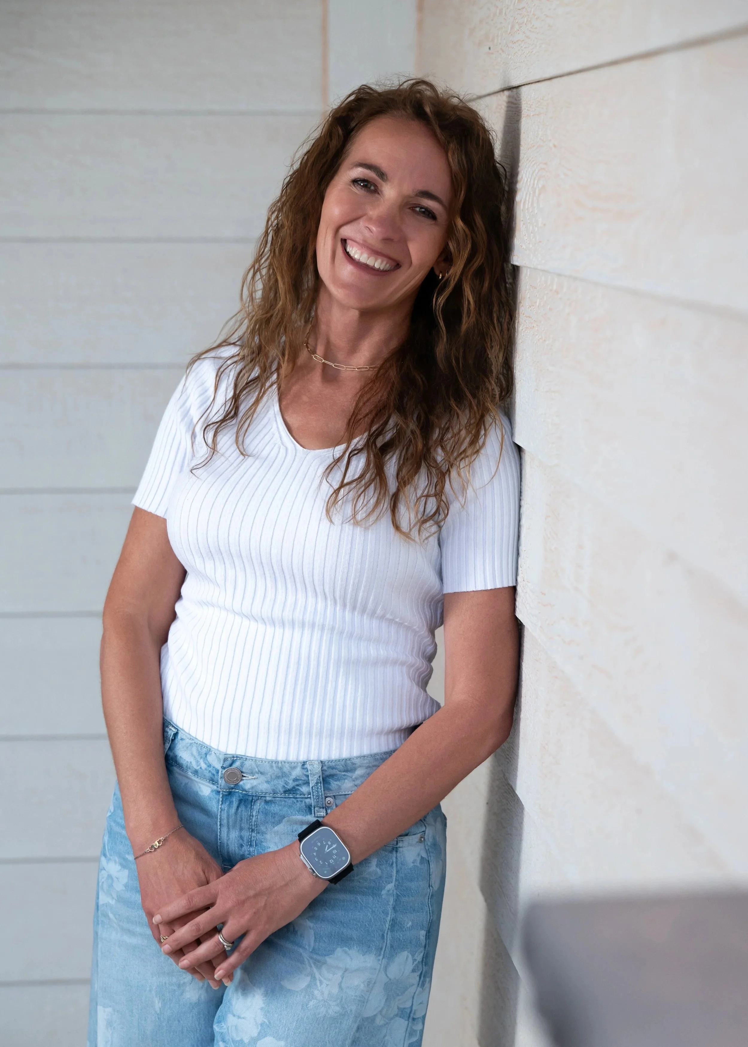 woman with curly brown hair smiling and leaning against a white wall, wearing a white ribbed shirt, blue denim jeans, a smartwatch, a chain necklace, and rings on her fingers.