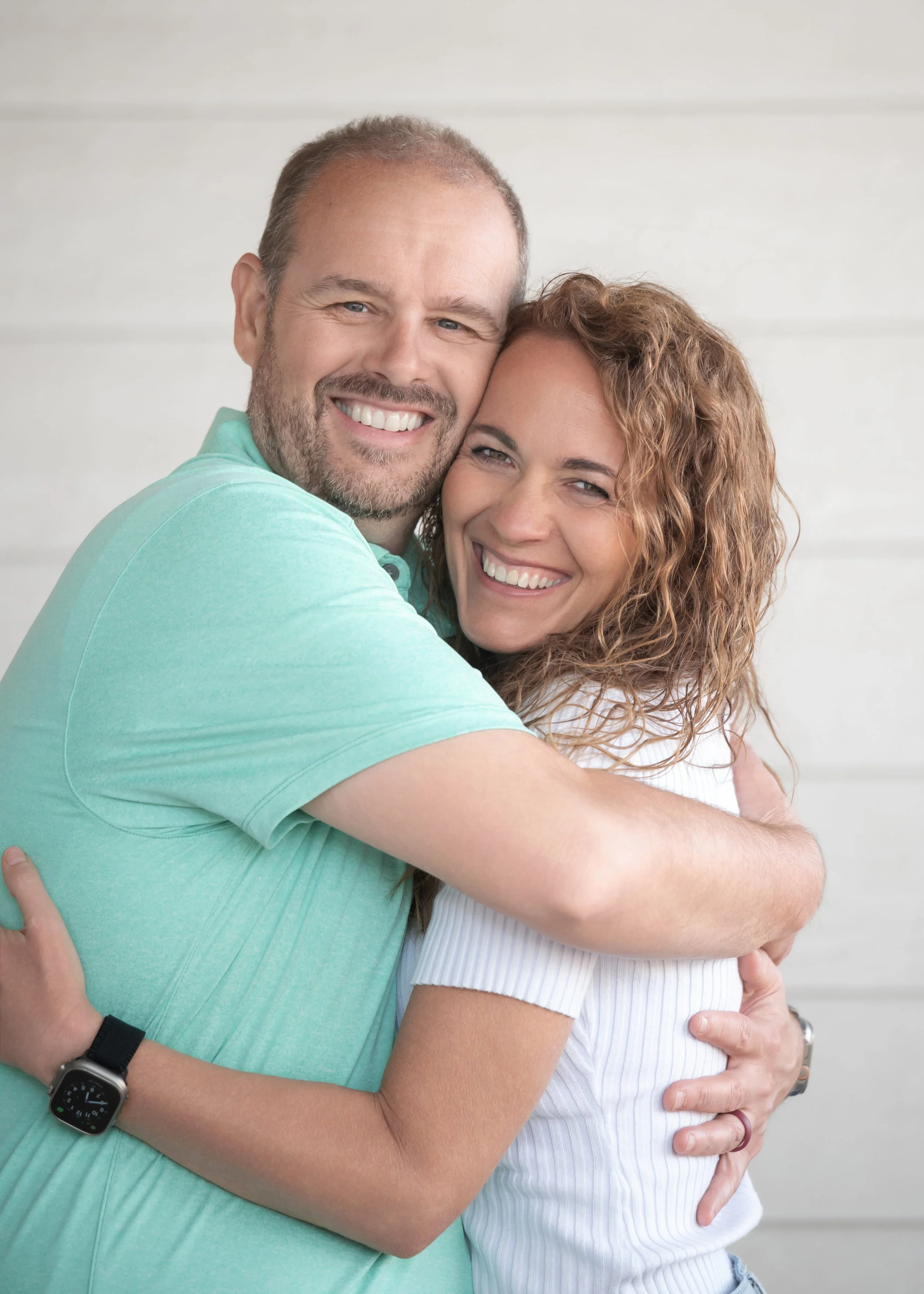 A happy couple hugging each other, smiling, with a white wall in the background.