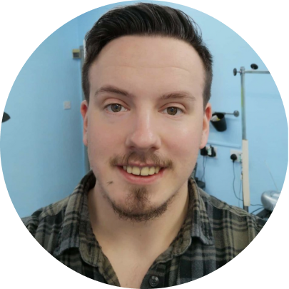 A young man with short dark hair, a goatee, and a mustache smiling in a room with light blue walls and fitness equipment.
