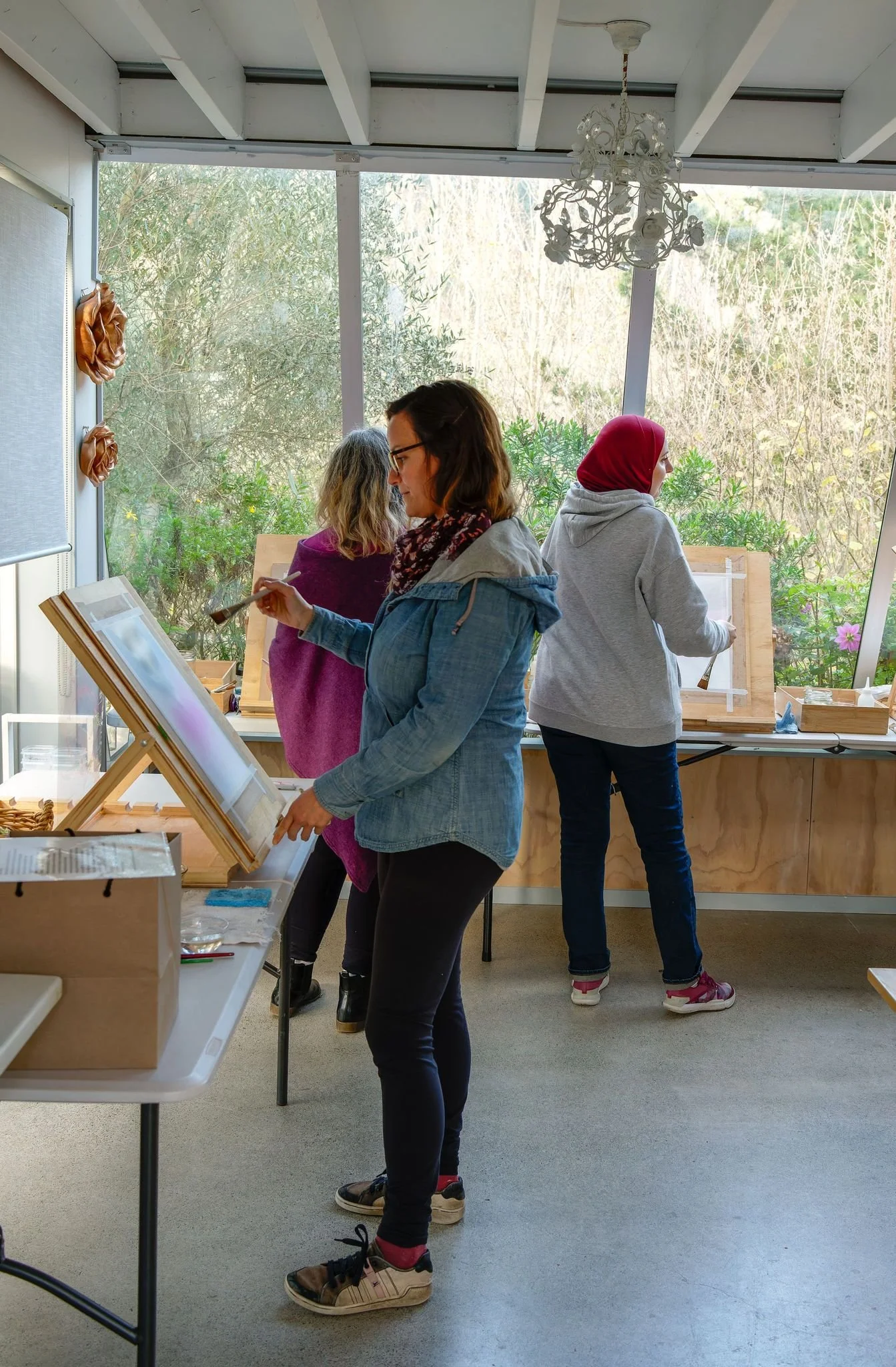 Three women painting at easels in a well-lit art studio with large windows and outdoor view.